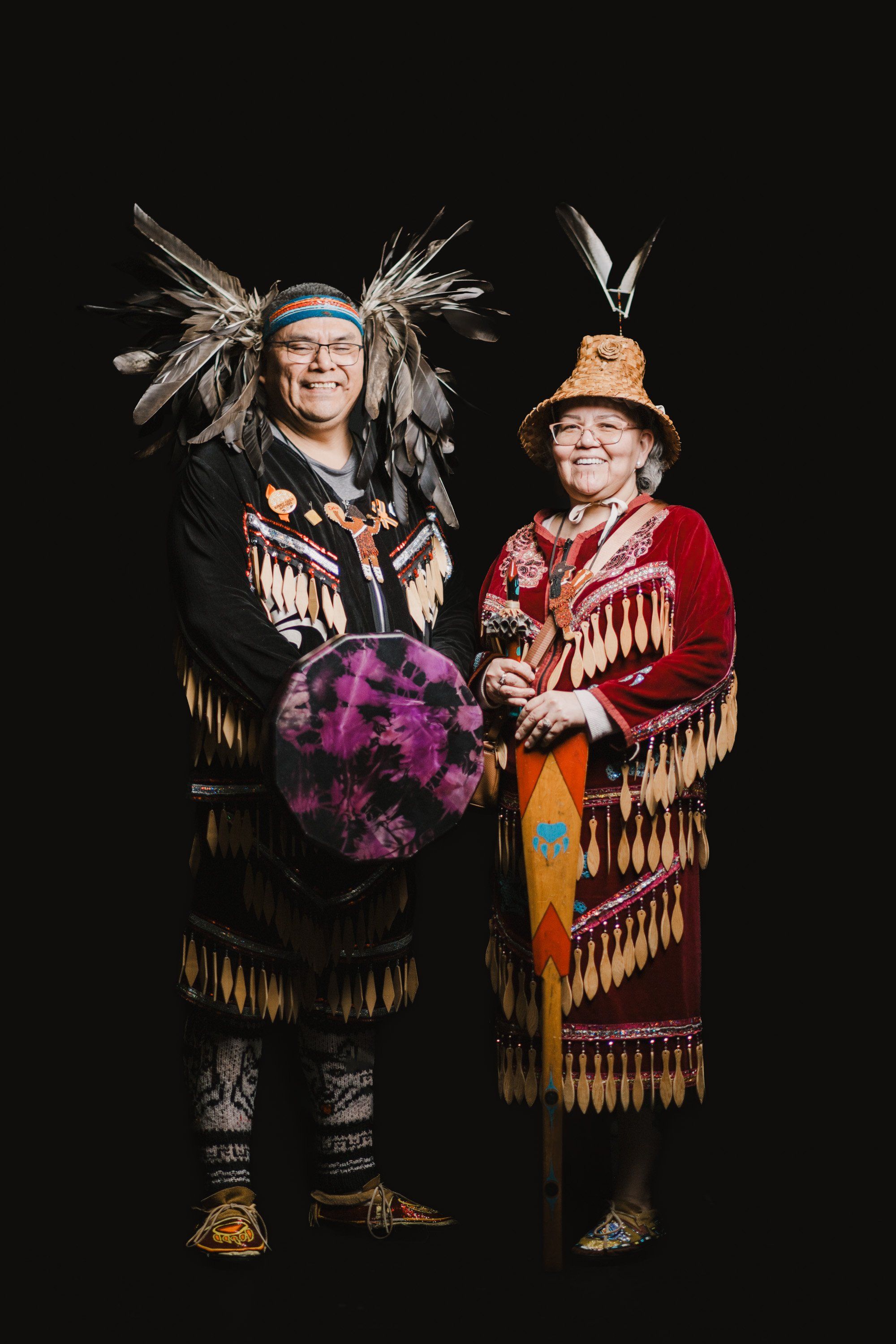 Two people in elaborate Coast Salish regalia with fringed button blankets and feathered headdresses smile warmly against a black background, holding a purple…