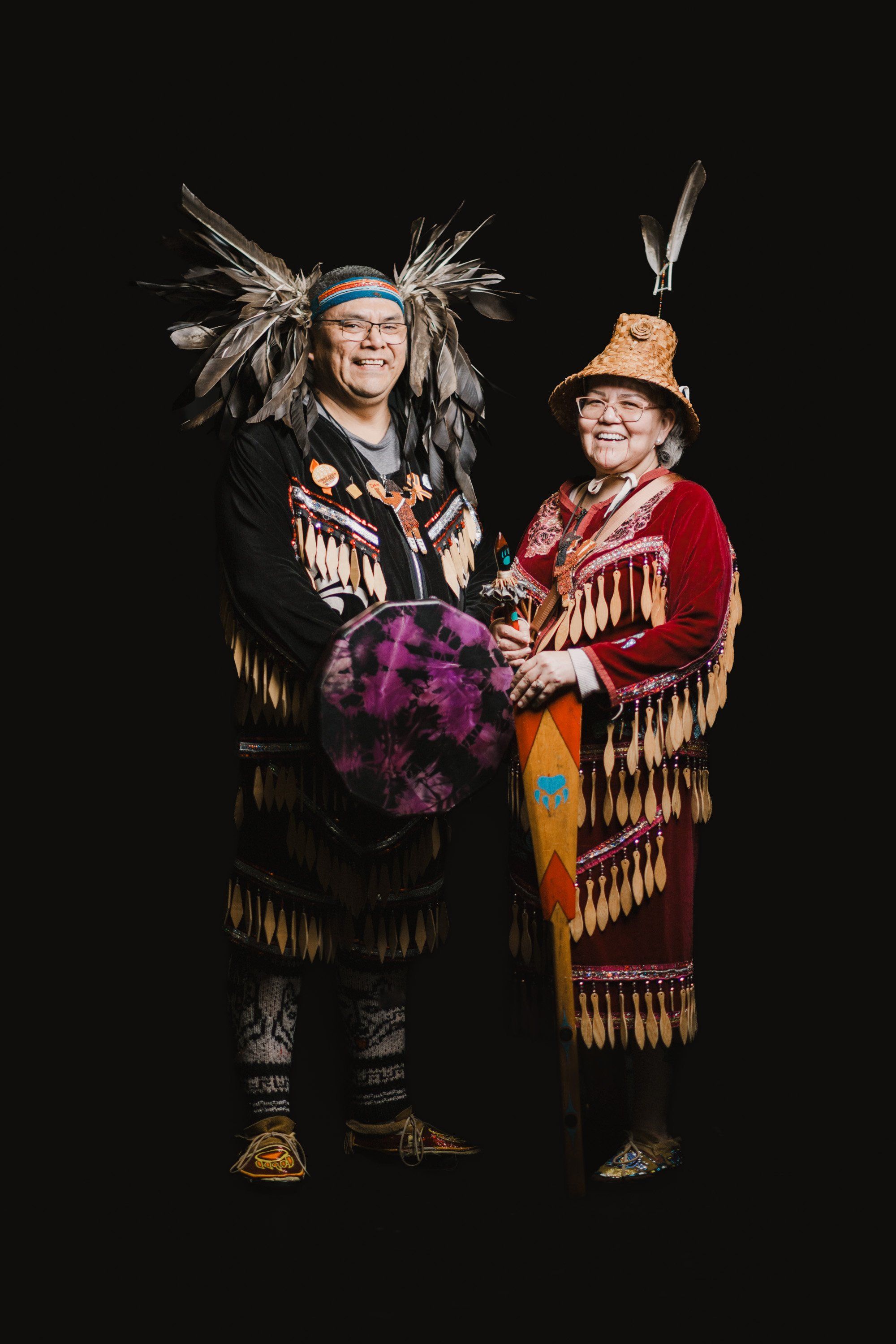 Two Coast Salish performers in traditional regalia with feathered headdresses hold a purple drum against a black background