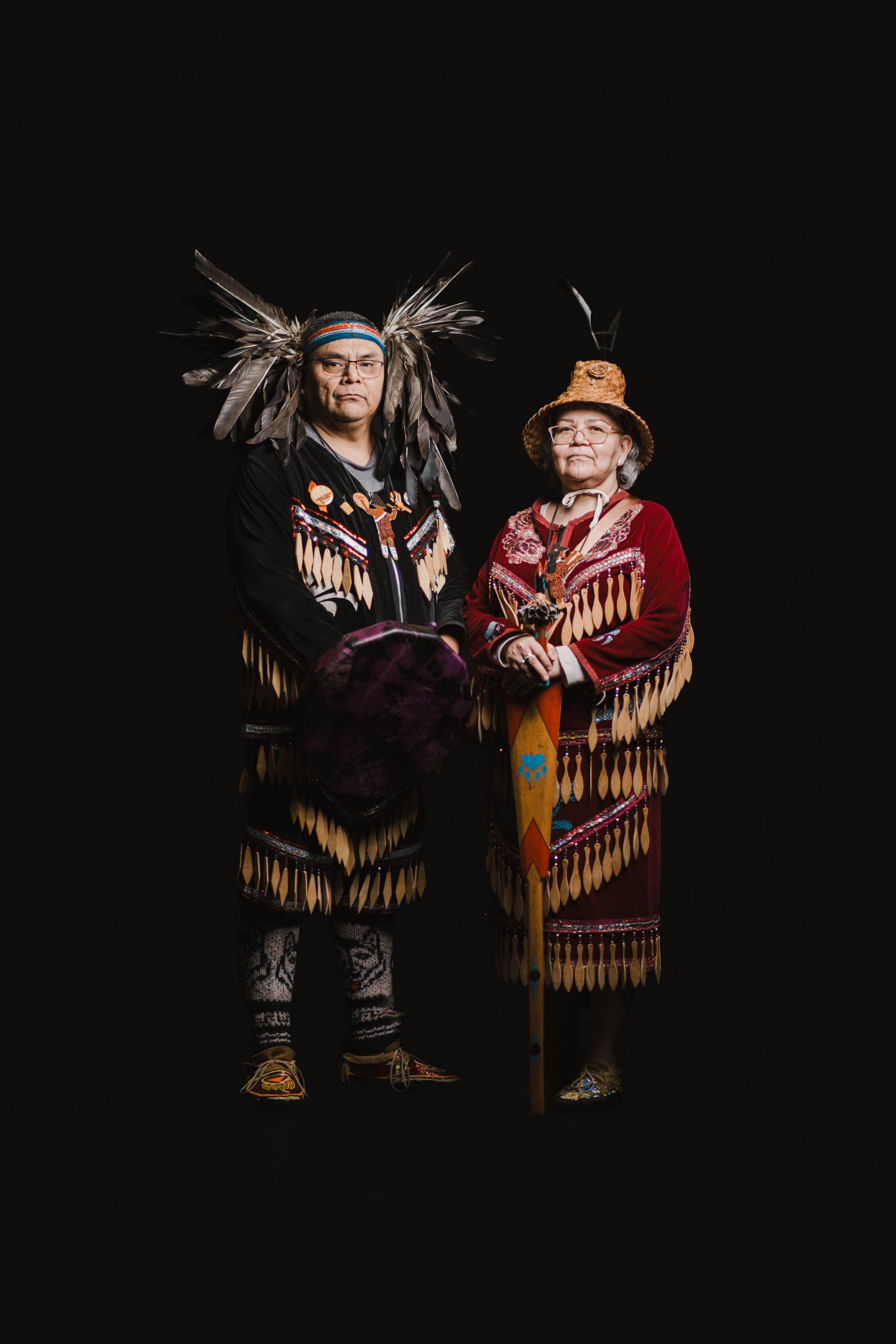 Two figures in elaborate traditional regalia with fringed button blankets and feathered headdresses stand together against black, their ornate regalia glowin…