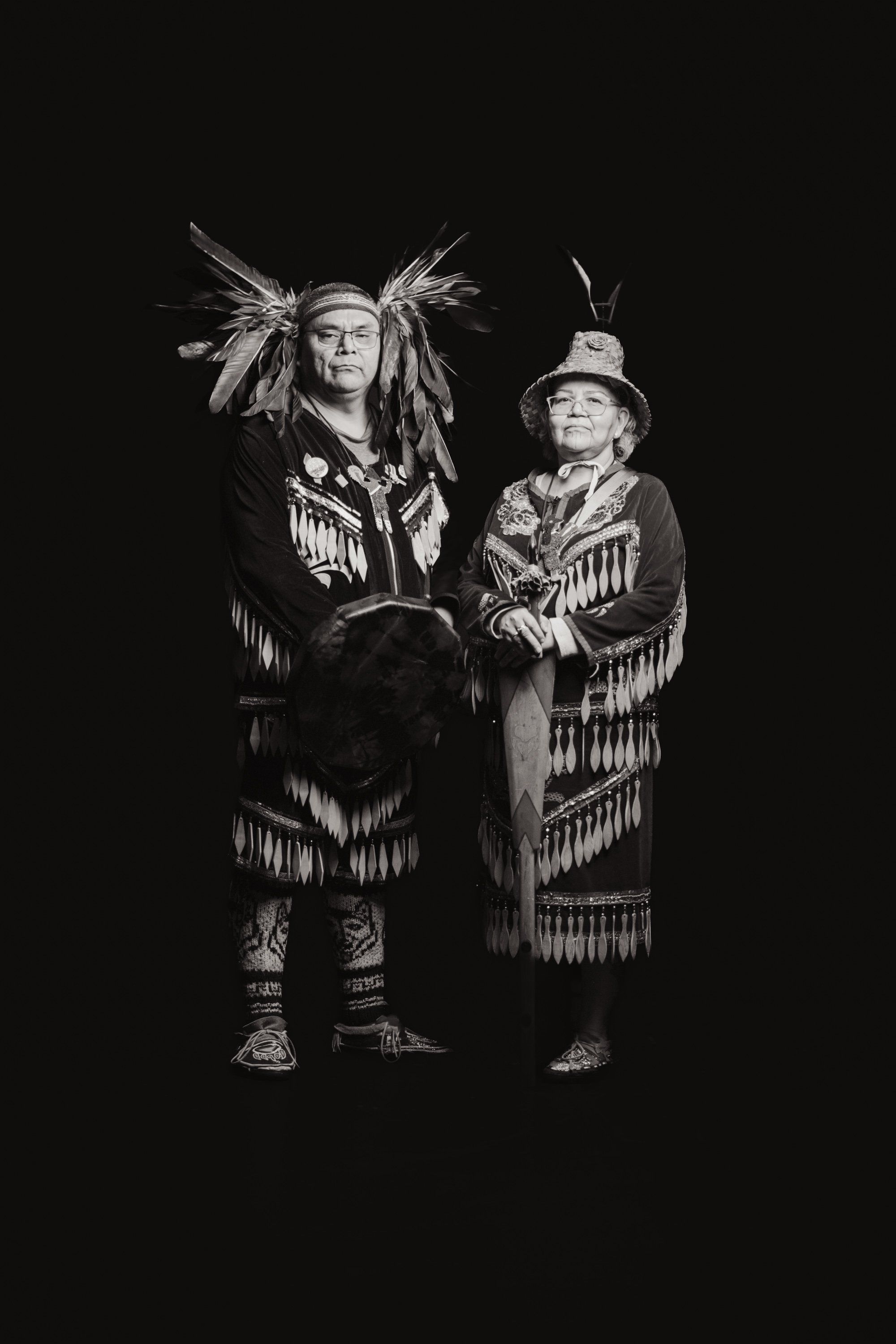 Two figures in traditional Coast Salish regalia stand against black, the taller wearing an elaborate headdress with feathers, both adorned with fringed blank…