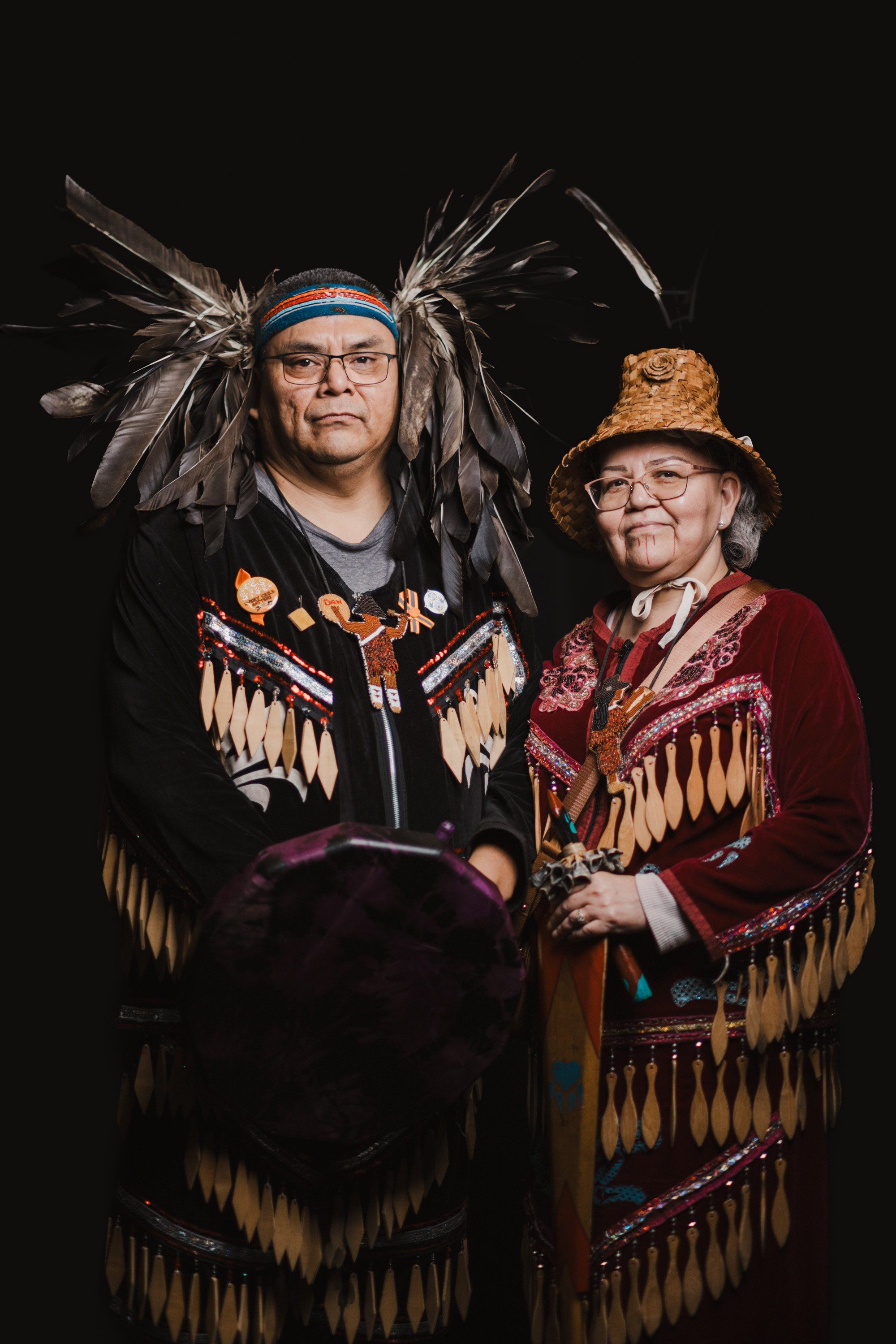 Two people in traditional Coast Salish regalia with fringed button blankets and headdresses stand together against a black background, smiling warmly at the…