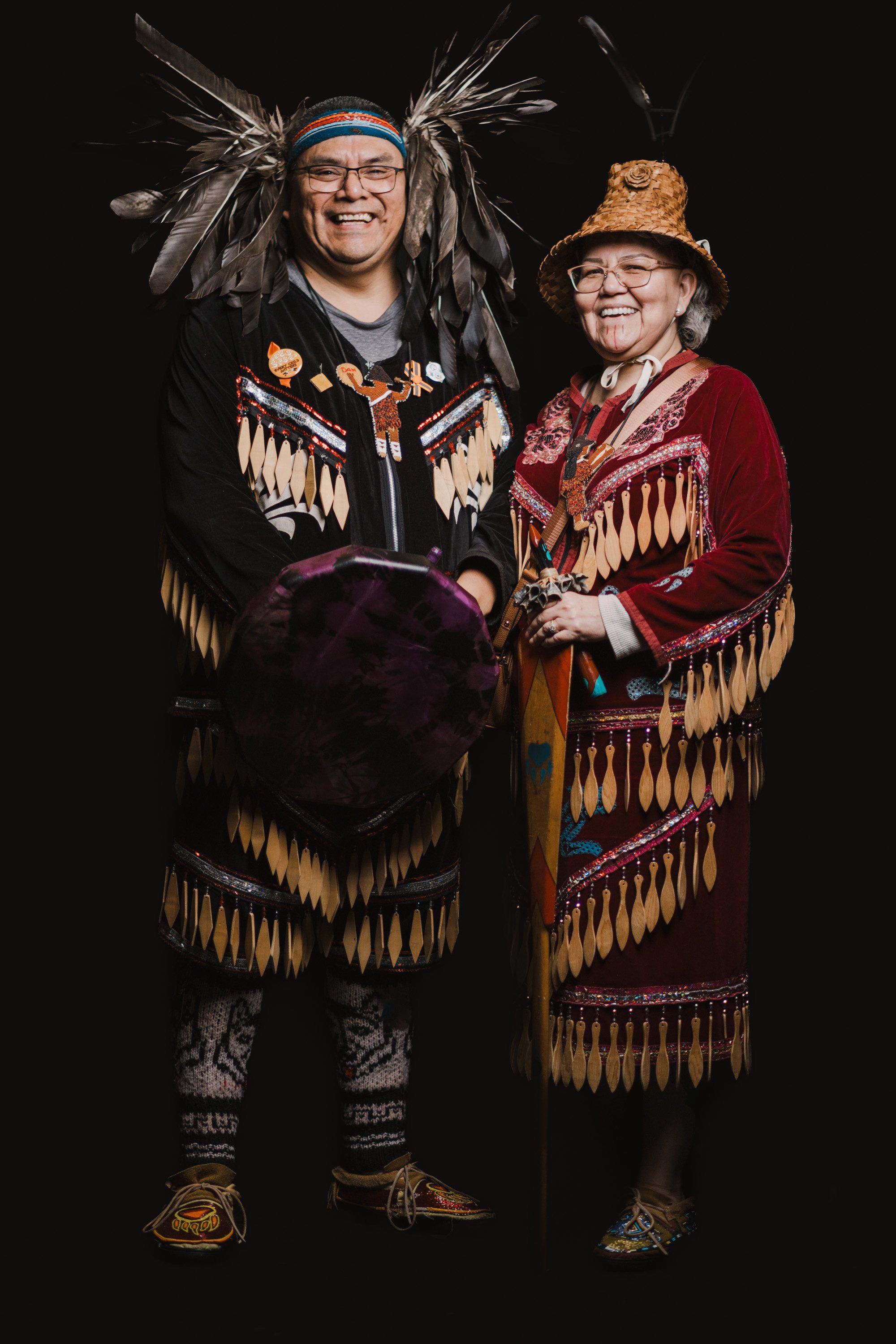 Two people in elaborate regalia with fringe and feathered headdresses stand smiling against a black background, celebrating Coast Salish culture and family