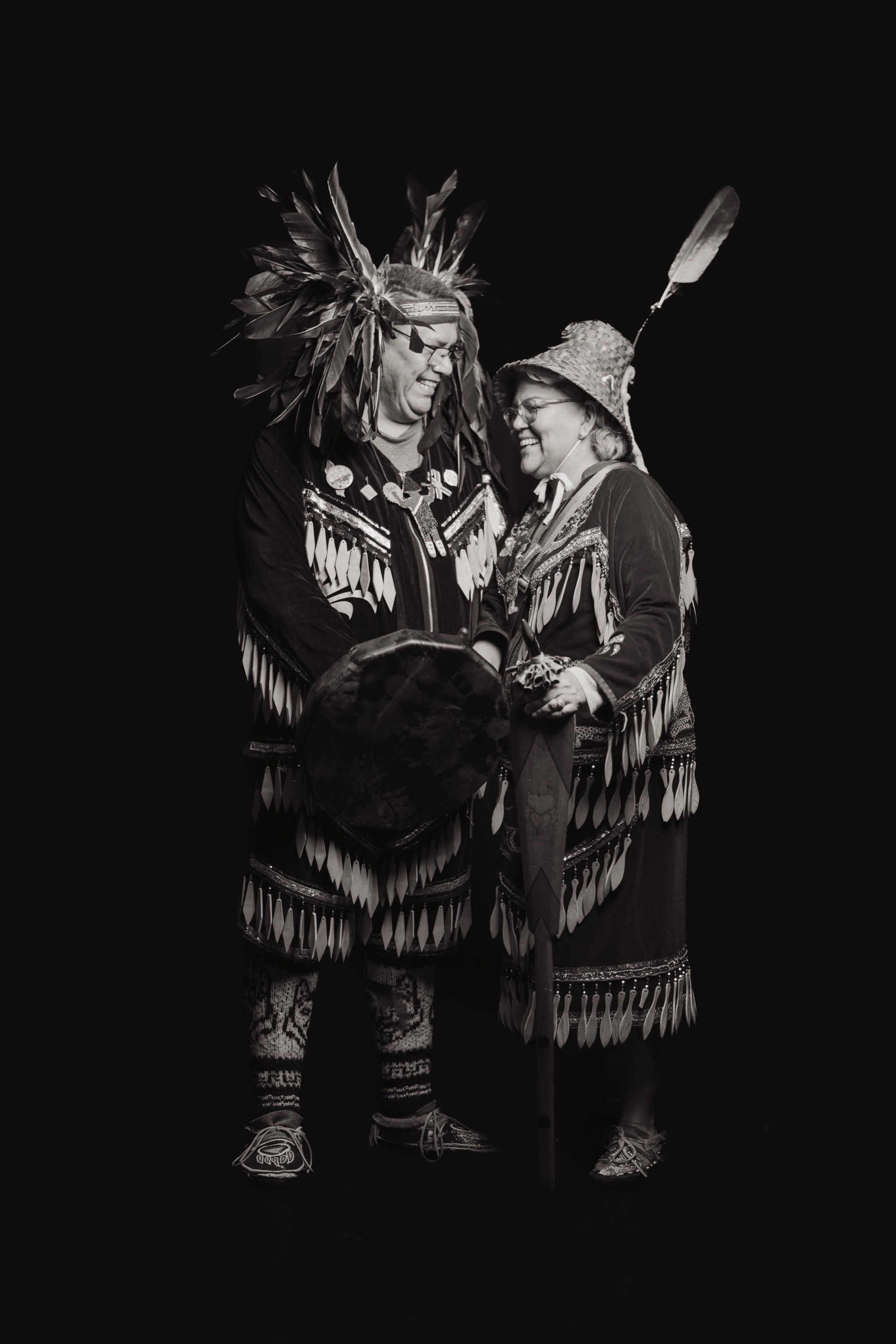 Two figures in traditional regalia with fringed clothing and headdresses stand together against black, sharing a moment of connection and joy