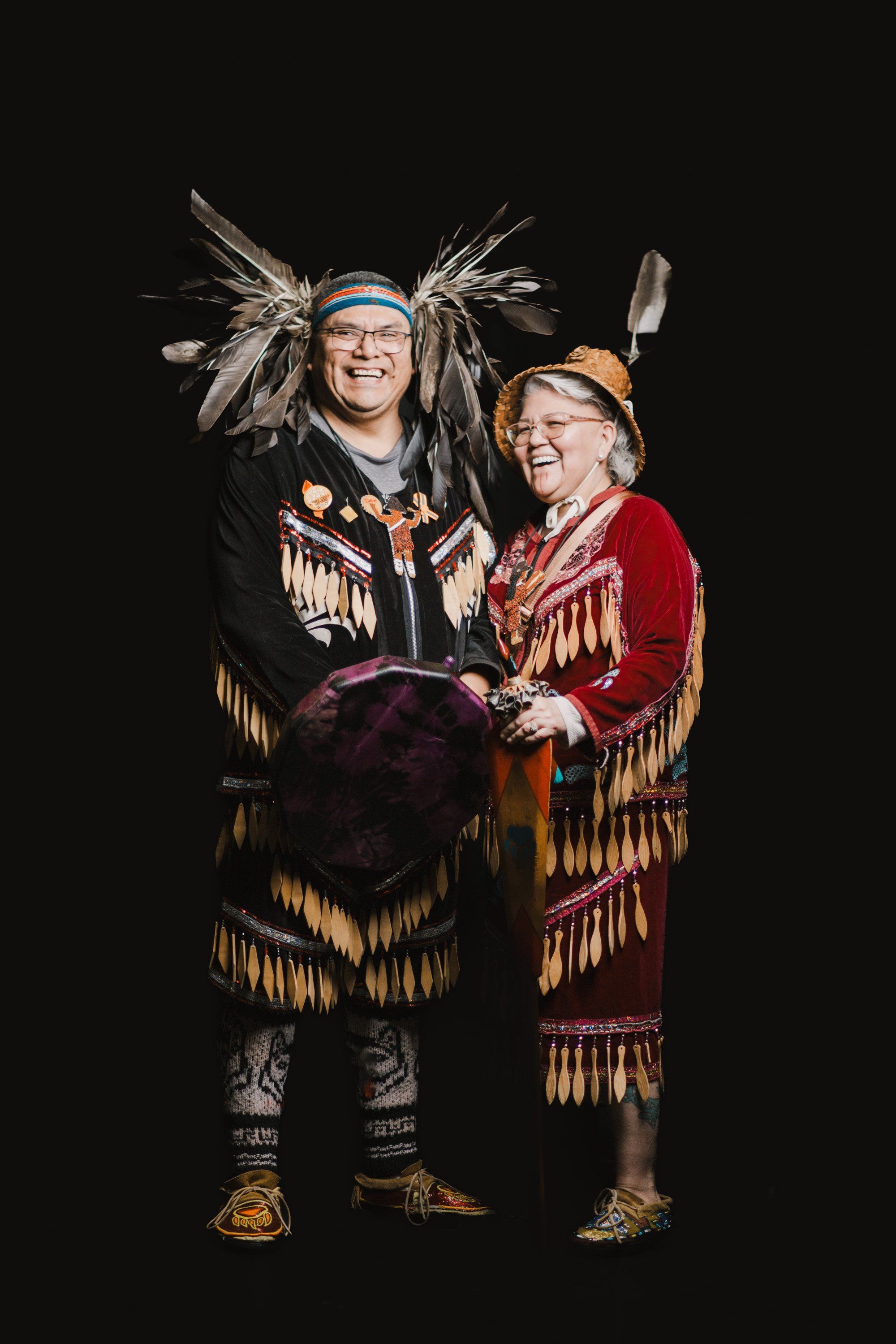 Two smiling people in elaborate fringed regalia and traditional headdresses pose together against a black background, holding a drum