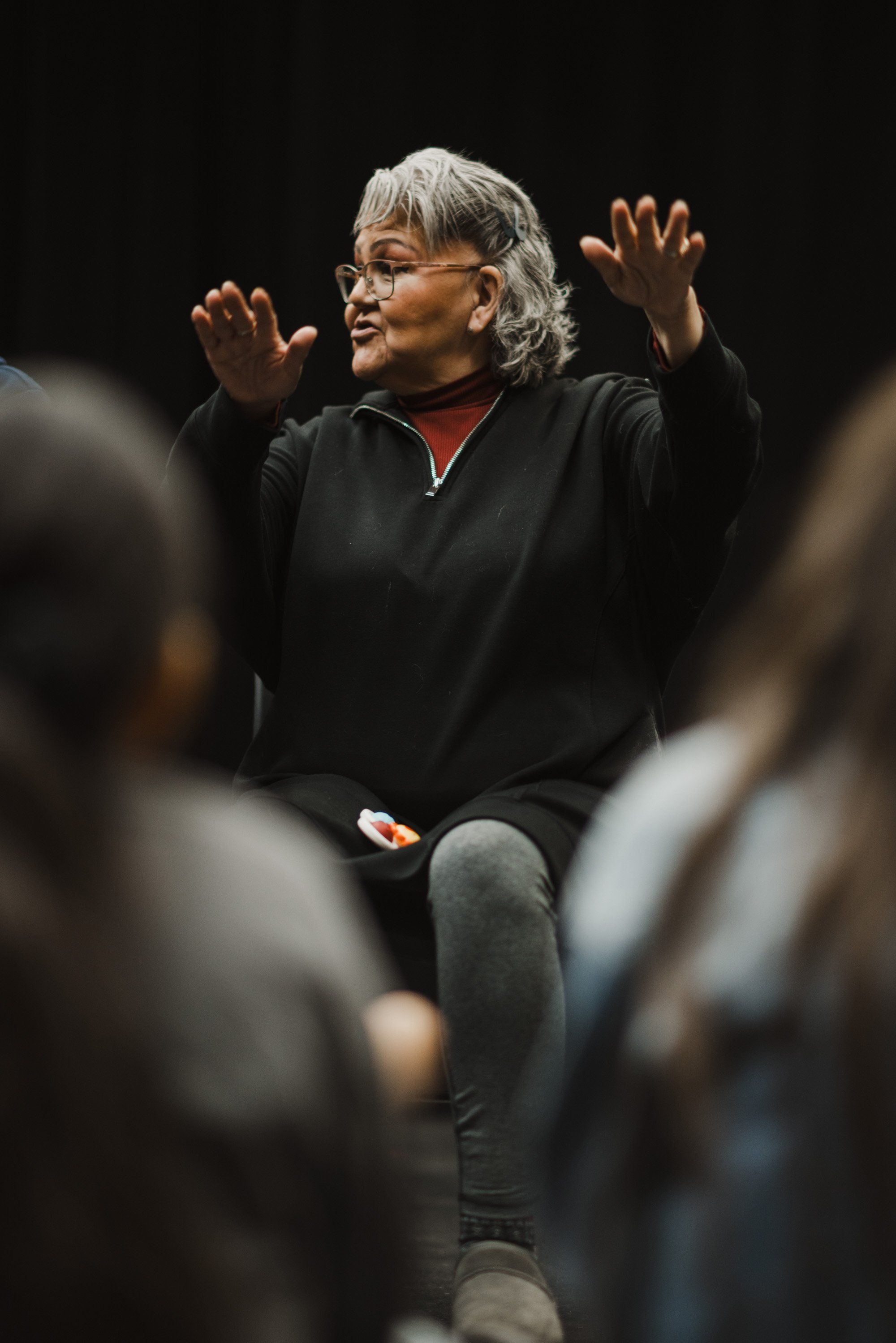 Woman with white hair and glasses gestures expressively while speaking, wearing black jacket over red shirt, with audience members blurred in foreground