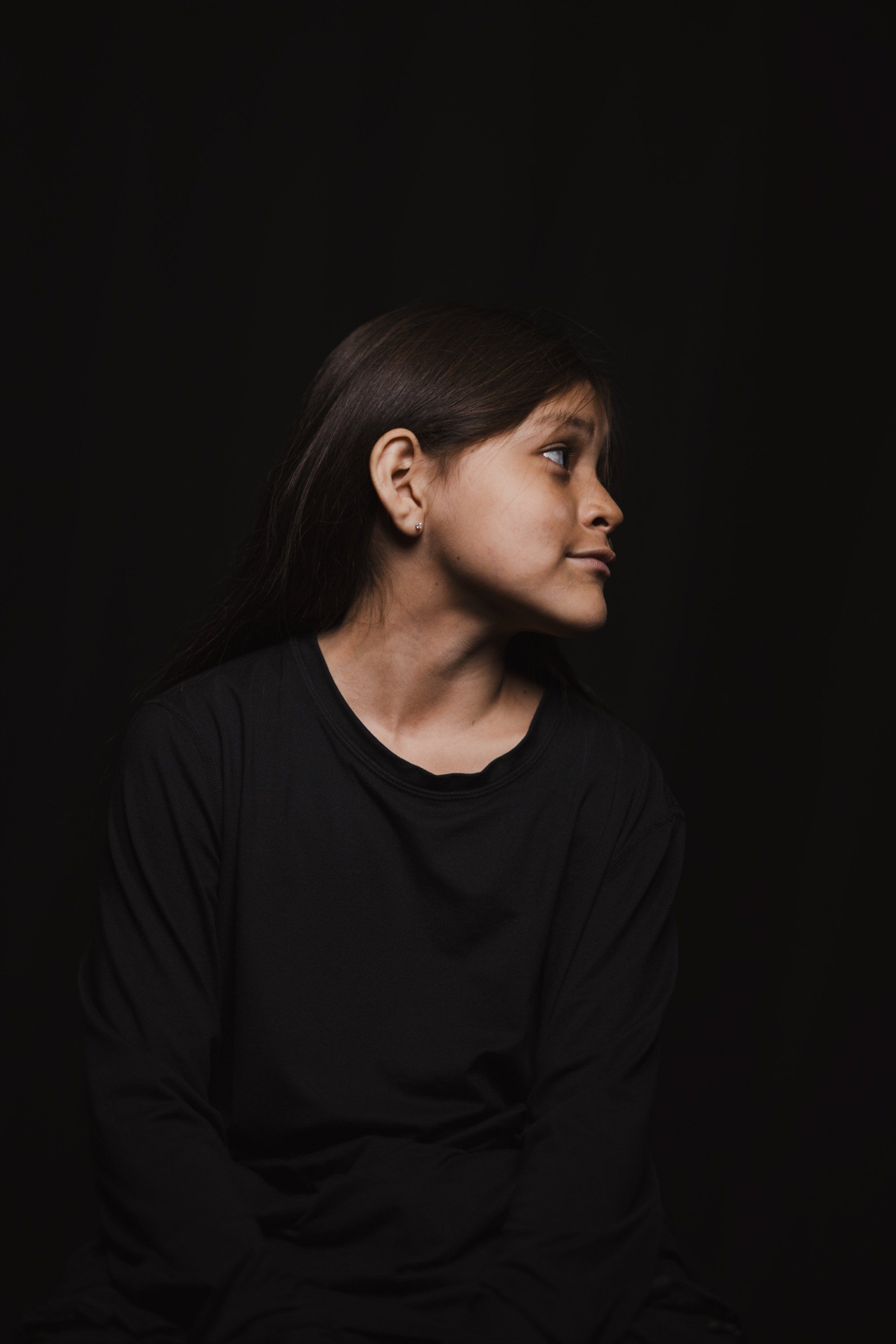 Woman in profile against black background, long dark hair, wearing black long-sleeved shirt, soft side lighting illuminating her face and shoulder