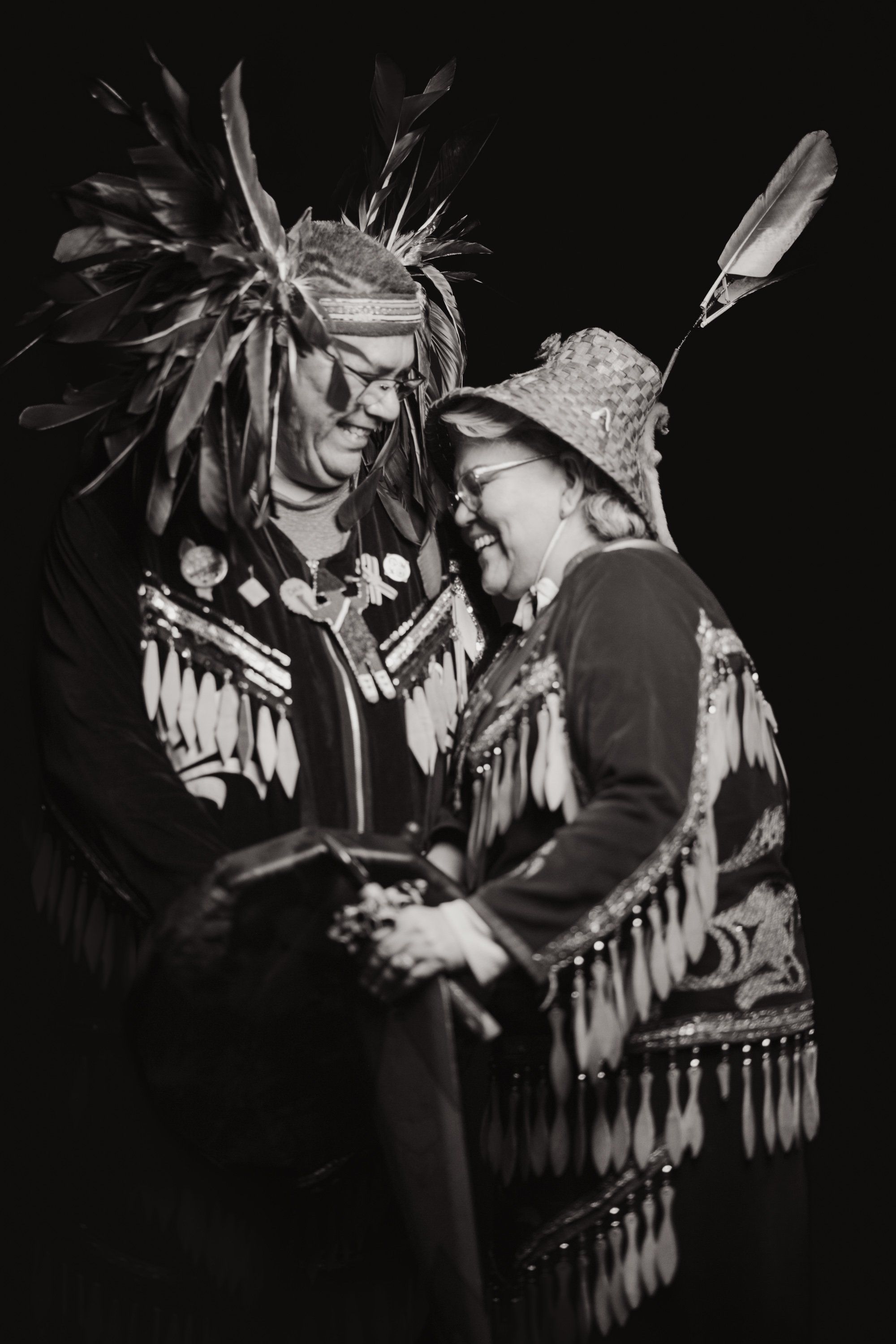 Two people in elaborate traditional regalia with feathered headdresses and fringed clothing embrace against a black background