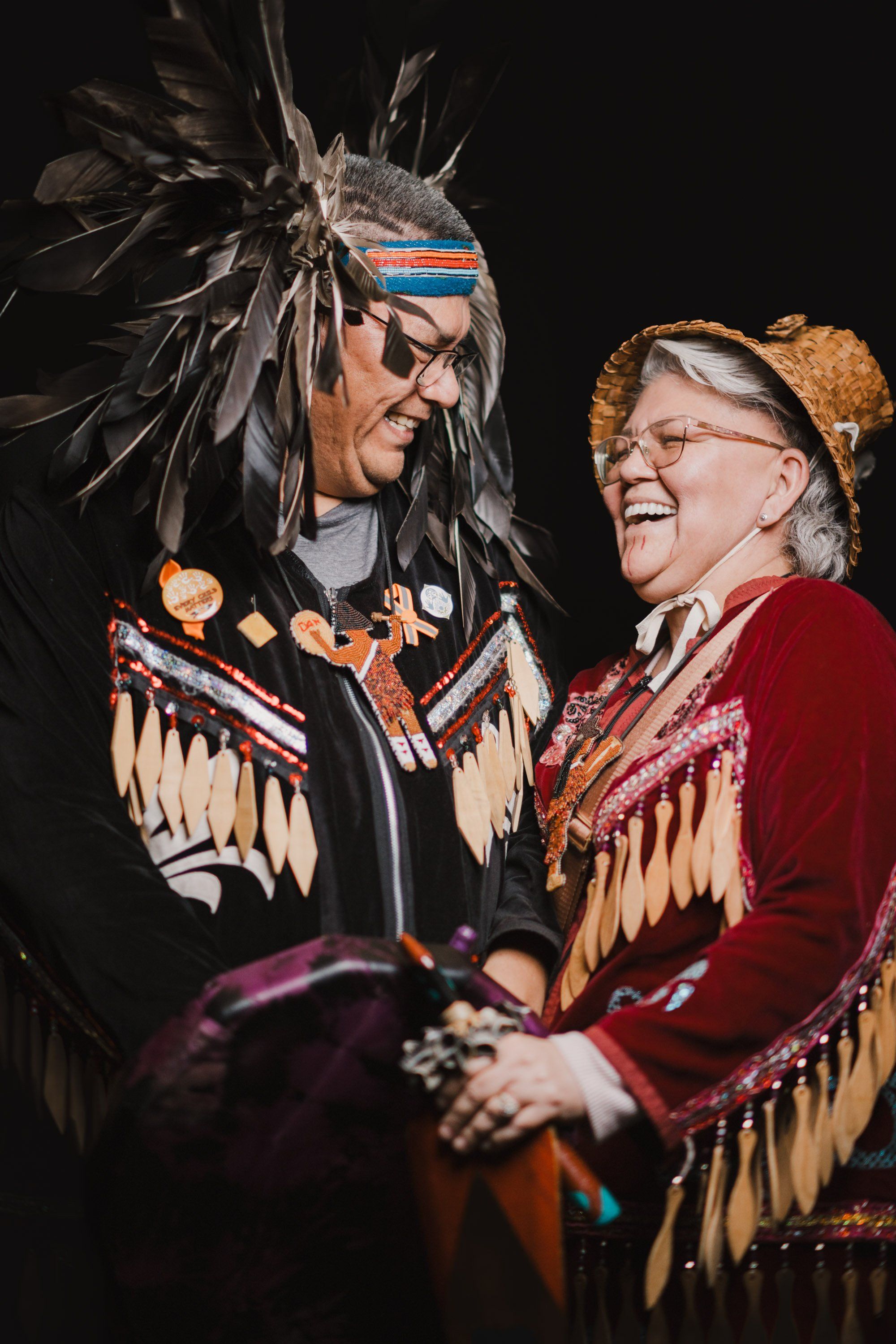 Two performers in traditional Coast Salish regalia smile warmly while holding hands against a black background, their ornate button blankets and headdresses…