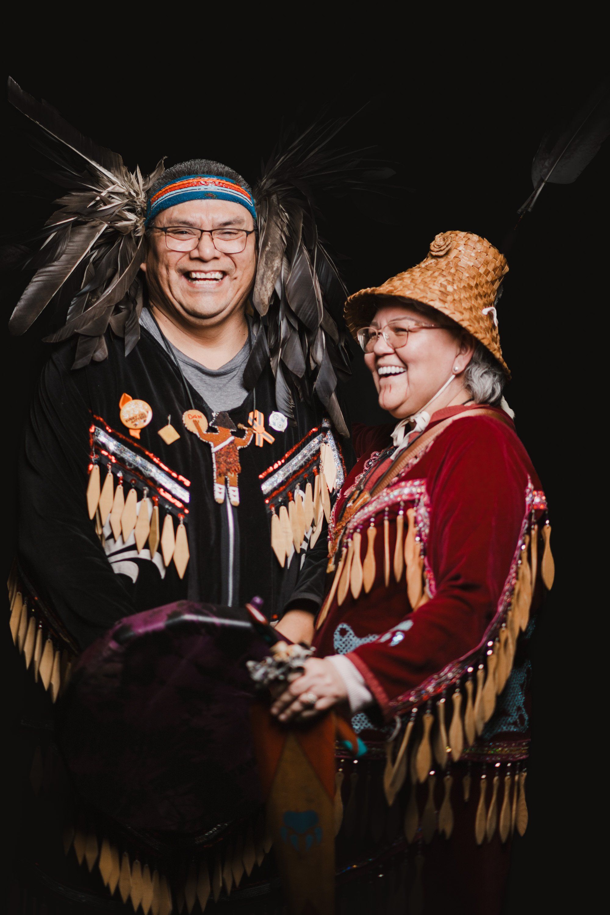 Two smiling performers in traditional Coast Salish regalia with fringed button blankets and feathered headdresses stand together against a black background