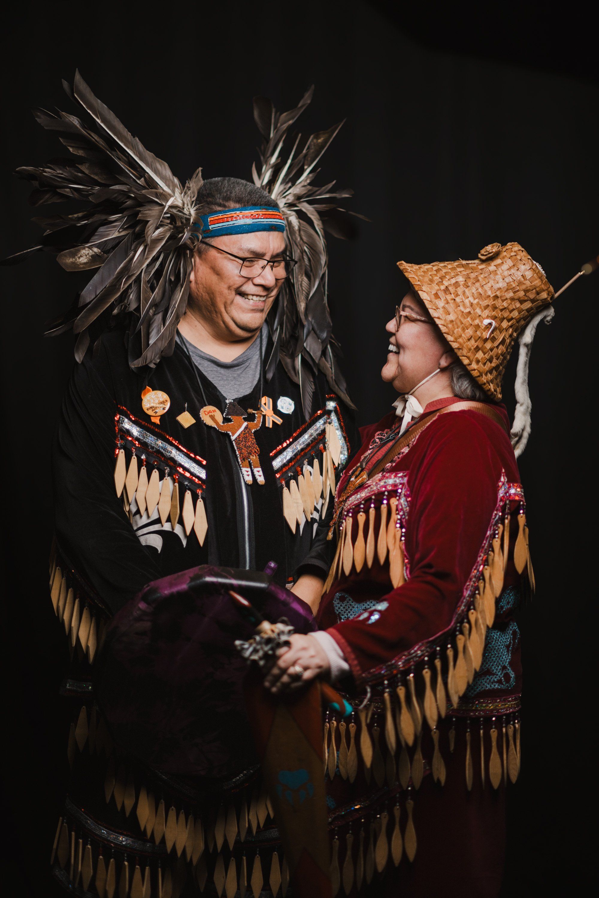 Two people in elaborate traditional Coast Salish regalia smile warmly at each other against a black background, their ornate button blankets and headdresses…