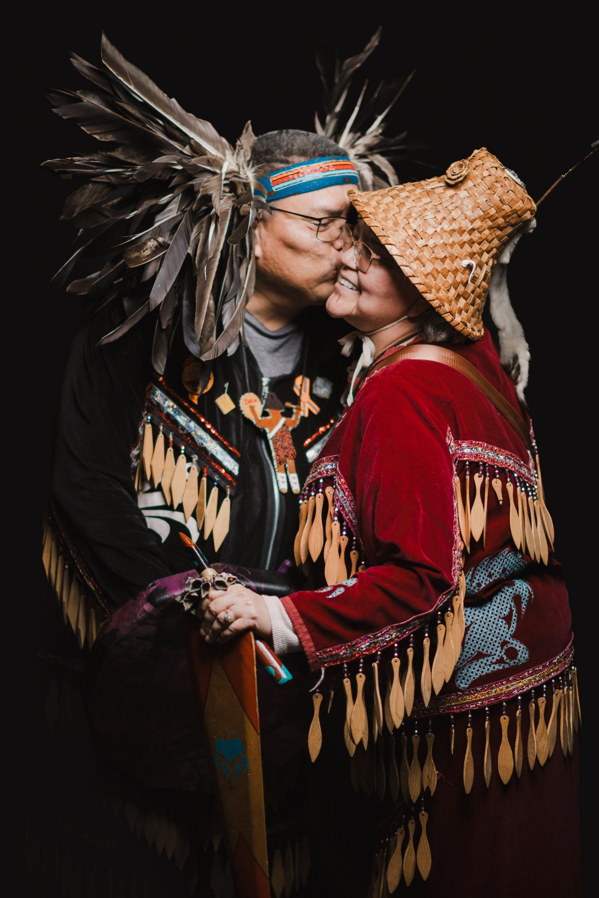 Two people in traditional Coast Salish regalia embrace against a black background, their ornate button blankets and headdresses adorned with feathers and geo…