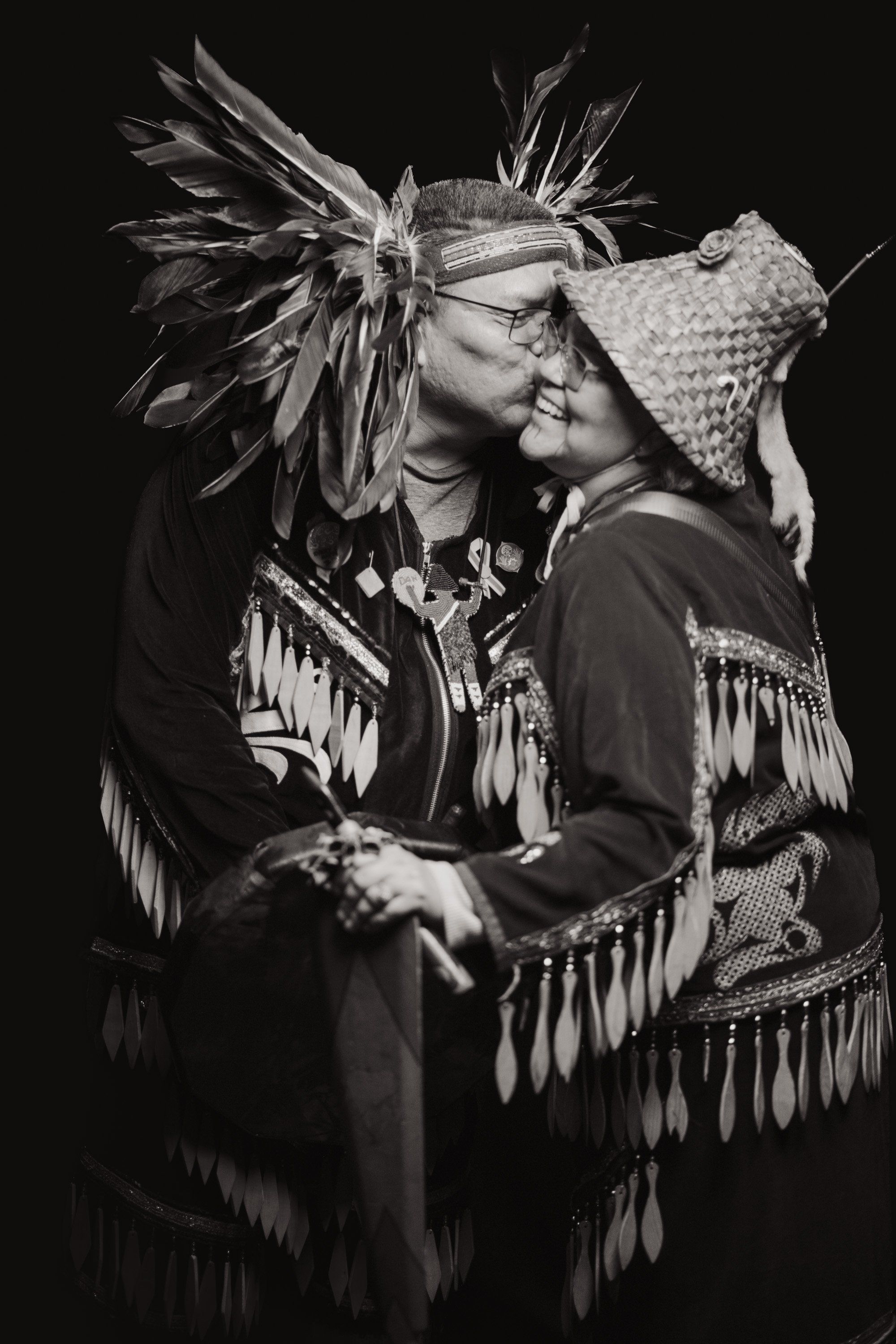 Two people in traditional Coast Salish regalia embrace in profile against black, their ornate button blankets and feathered headdress catching dramatic side…