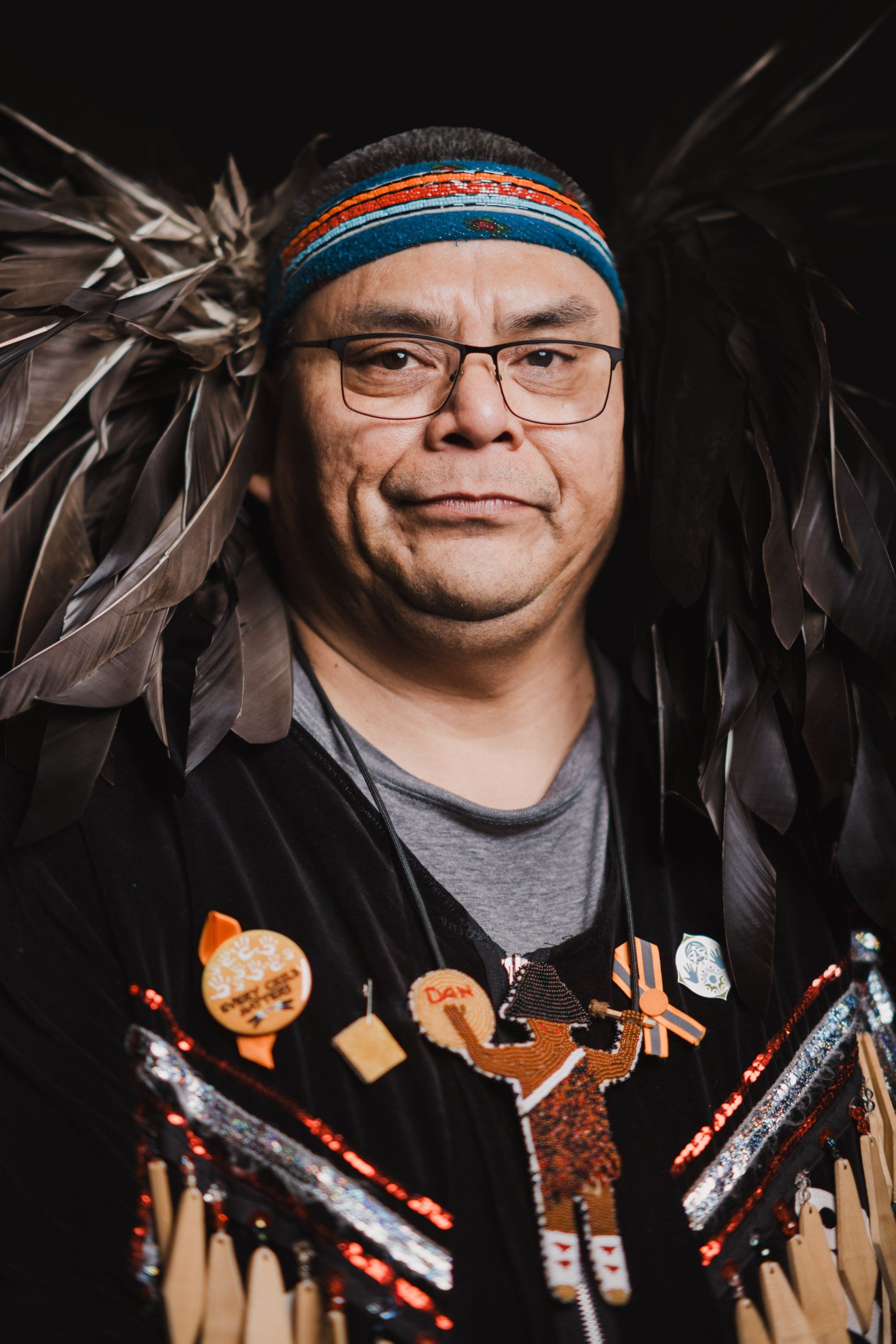 Portrait of a man wearing glasses and a blue and orange headband, adorned in traditional regalia with feather details, against a dark background