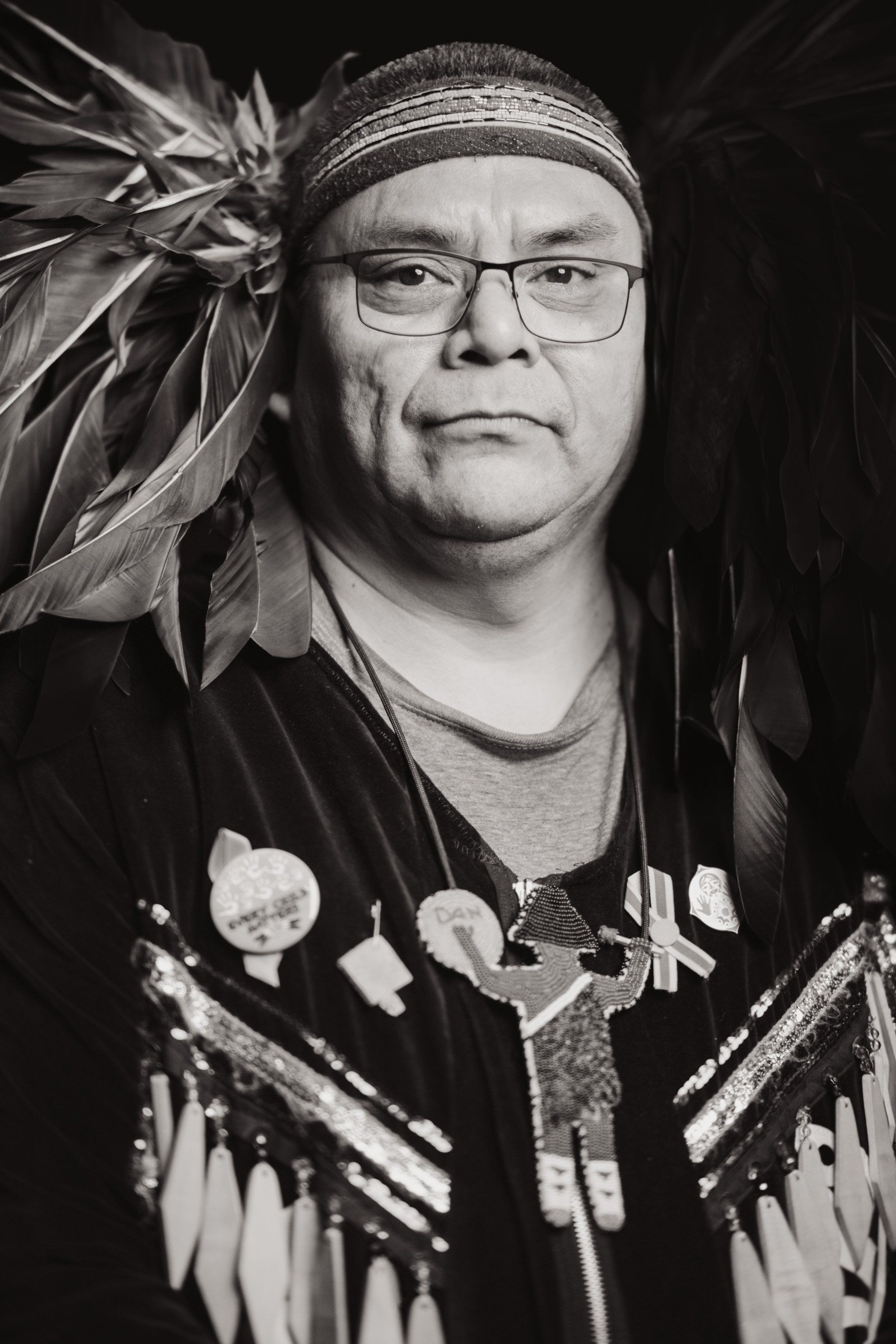 Portrait of a man wearing glasses and a headband, dressed in traditional Coast Salish regalia with feathered headdress and beaded vest, photographed against…
