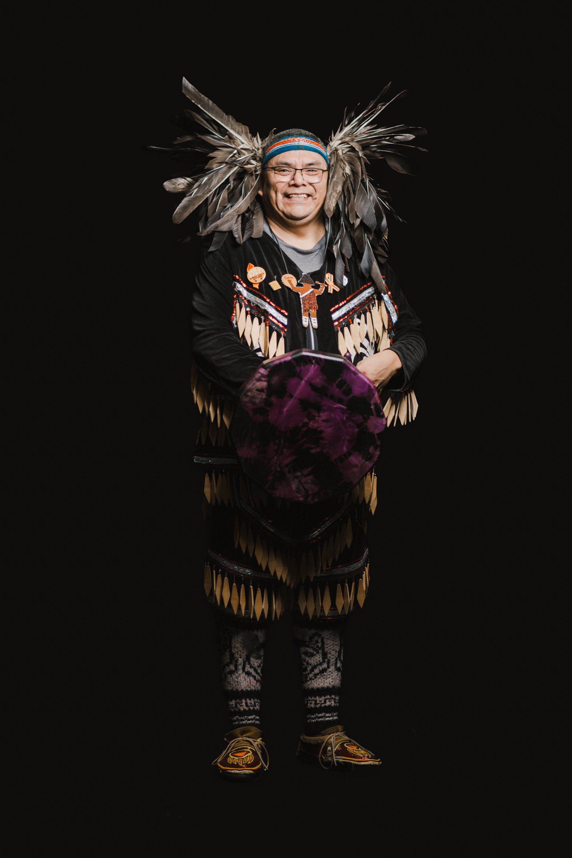 Smiling person wearing traditional Coast Salish regalia with feathered headdress, fringed leather vest, and purple skirt against black background