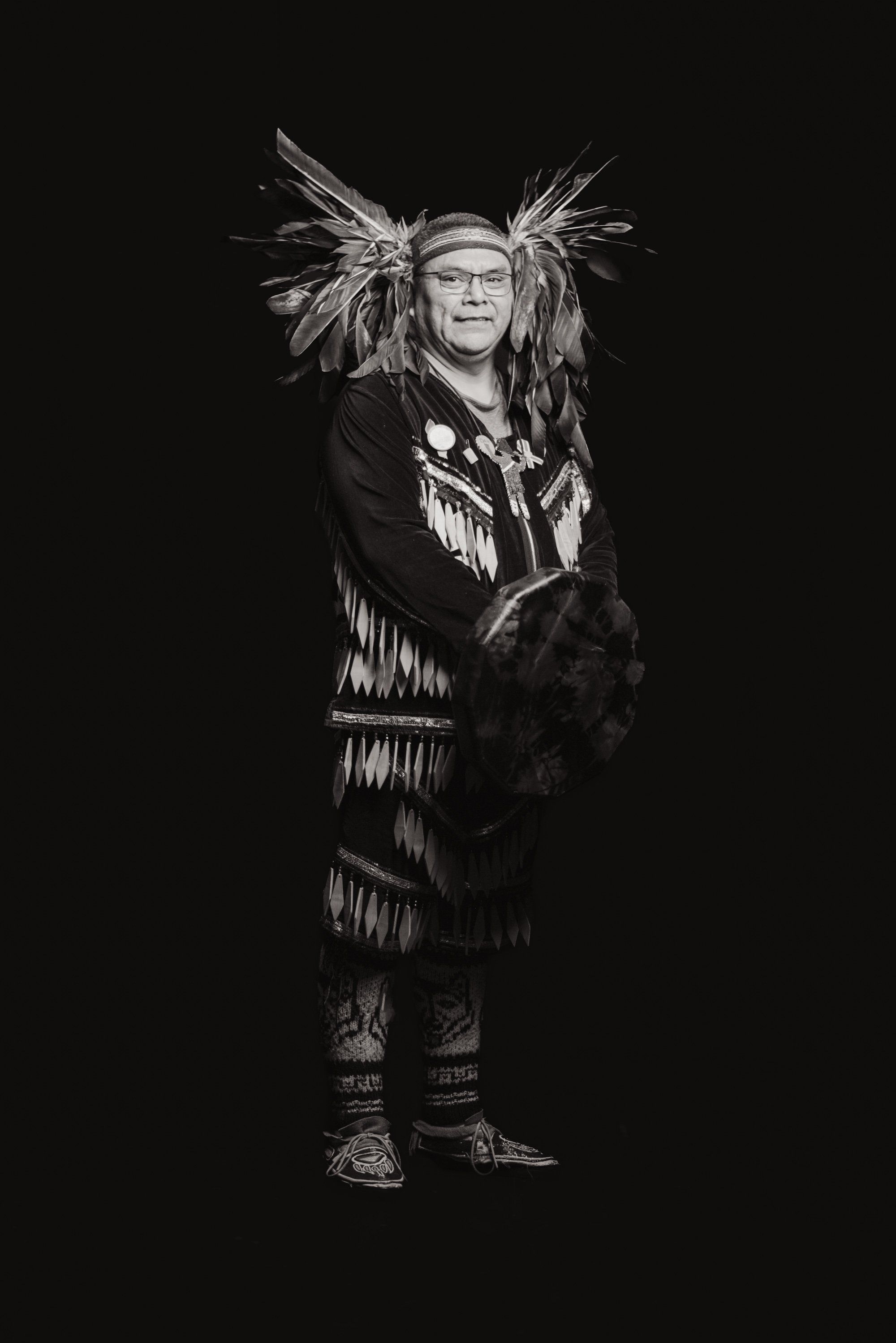 Man in traditional Coast Salish regalia with feathered headdress and fringed button blanket stands against black background, smiling at camera