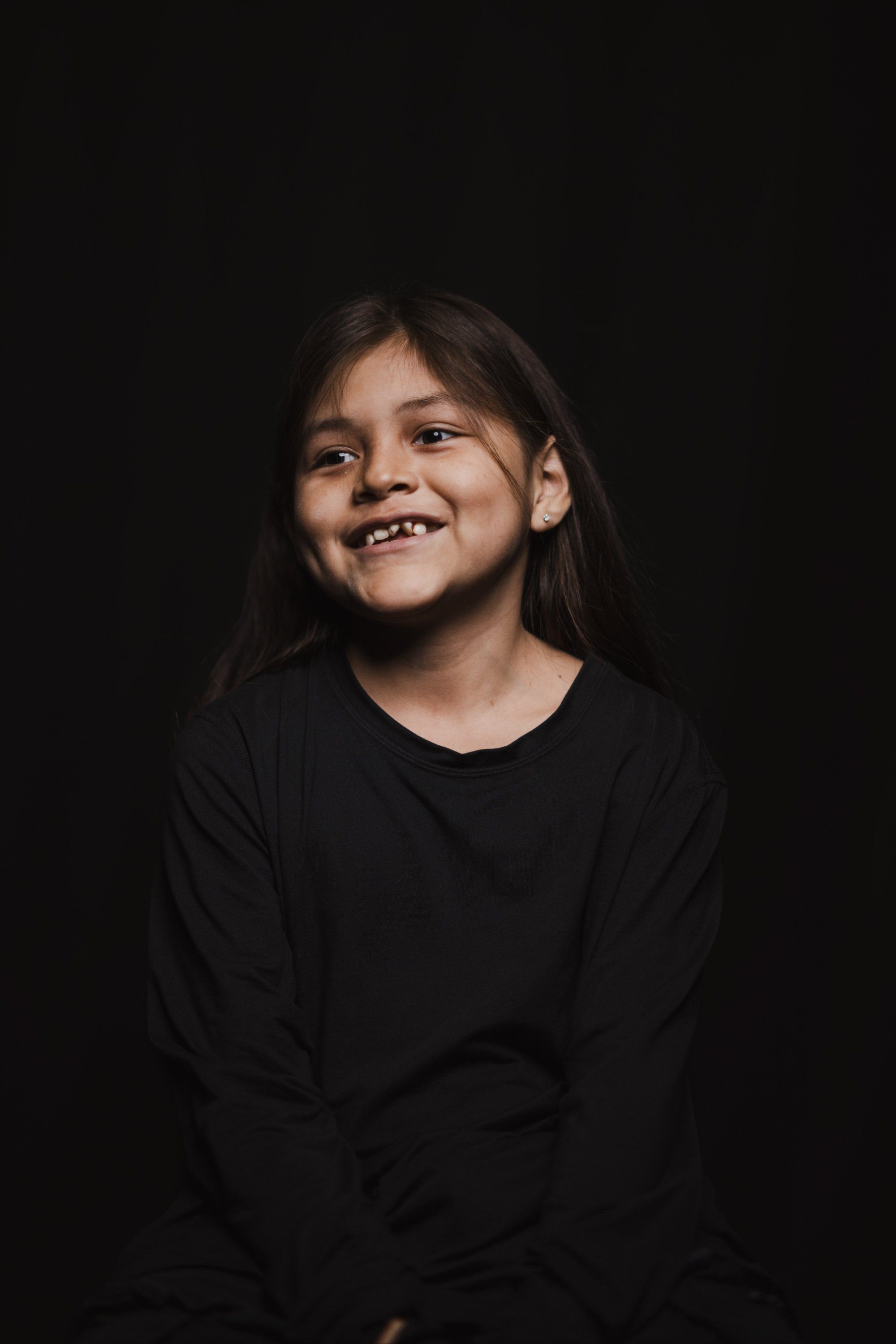 Young girl with long dark hair smiling warmly against a black background, wearing a black long-sleeved shirt