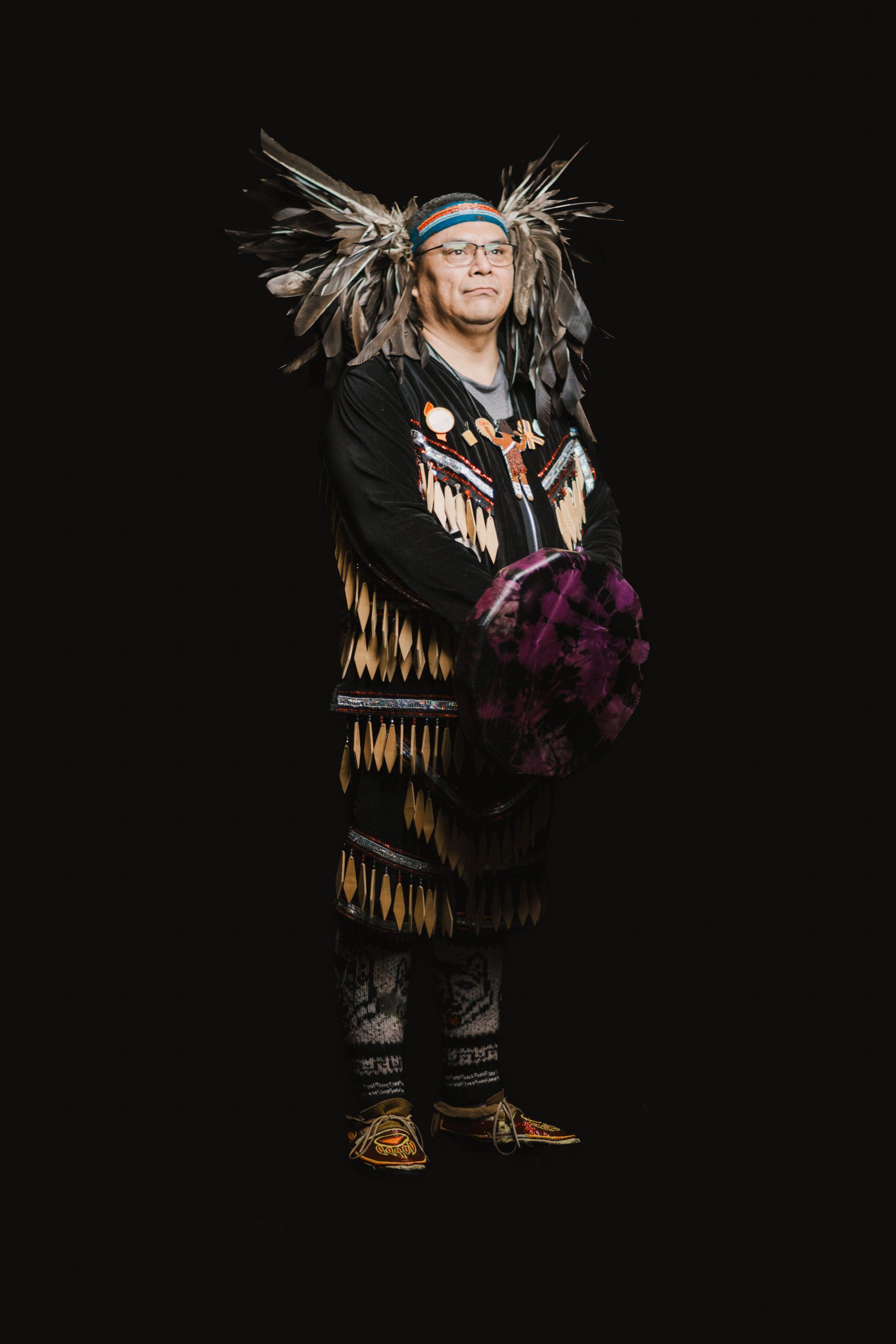 Elder in traditional Coast Salish regalia with feathered headdress and fringed button blanket stands against black background, holding purple fabric