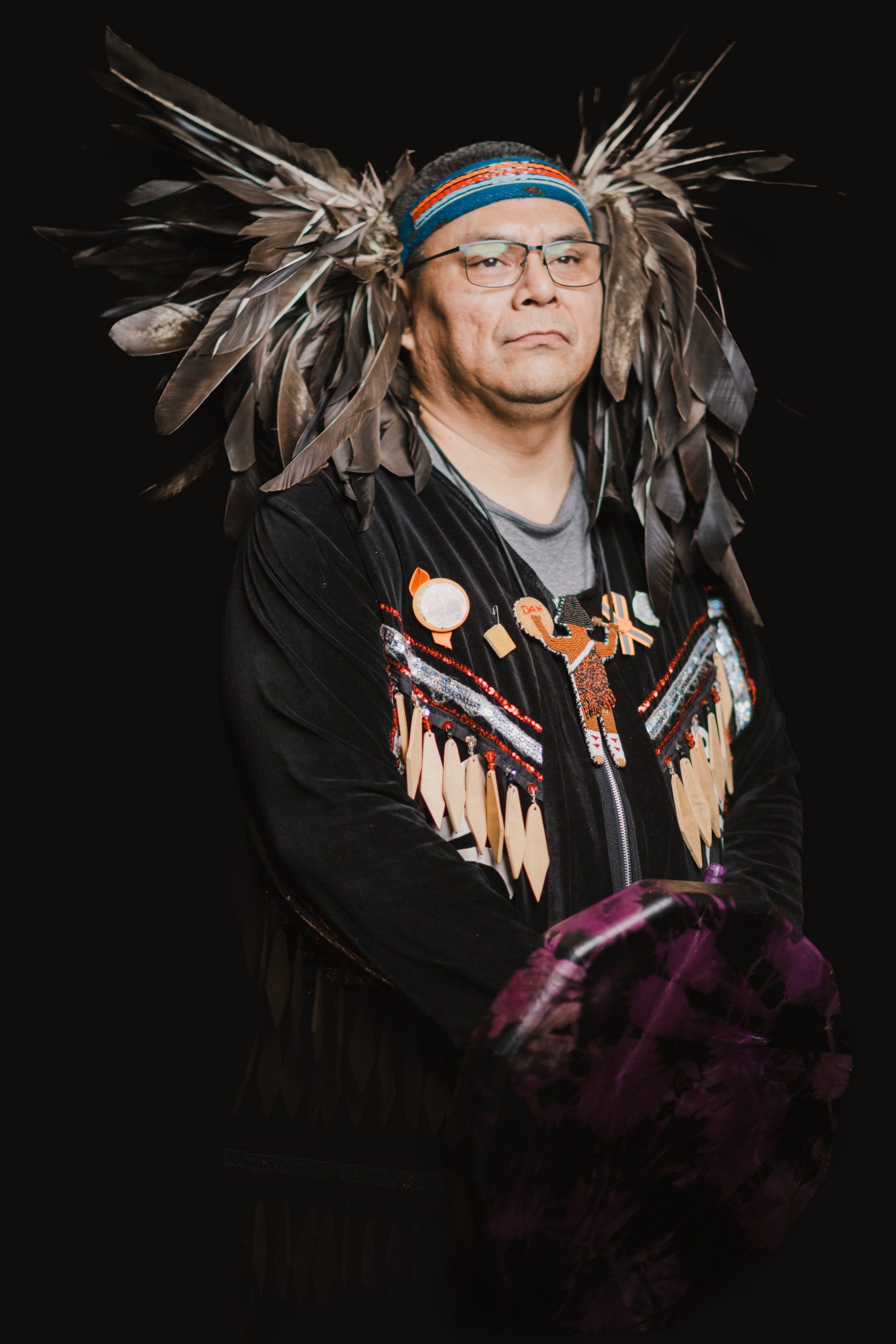 Coast Salish dancer in traditional regalia with feathered headdress and fringed vest gazes thoughtfully against dark background