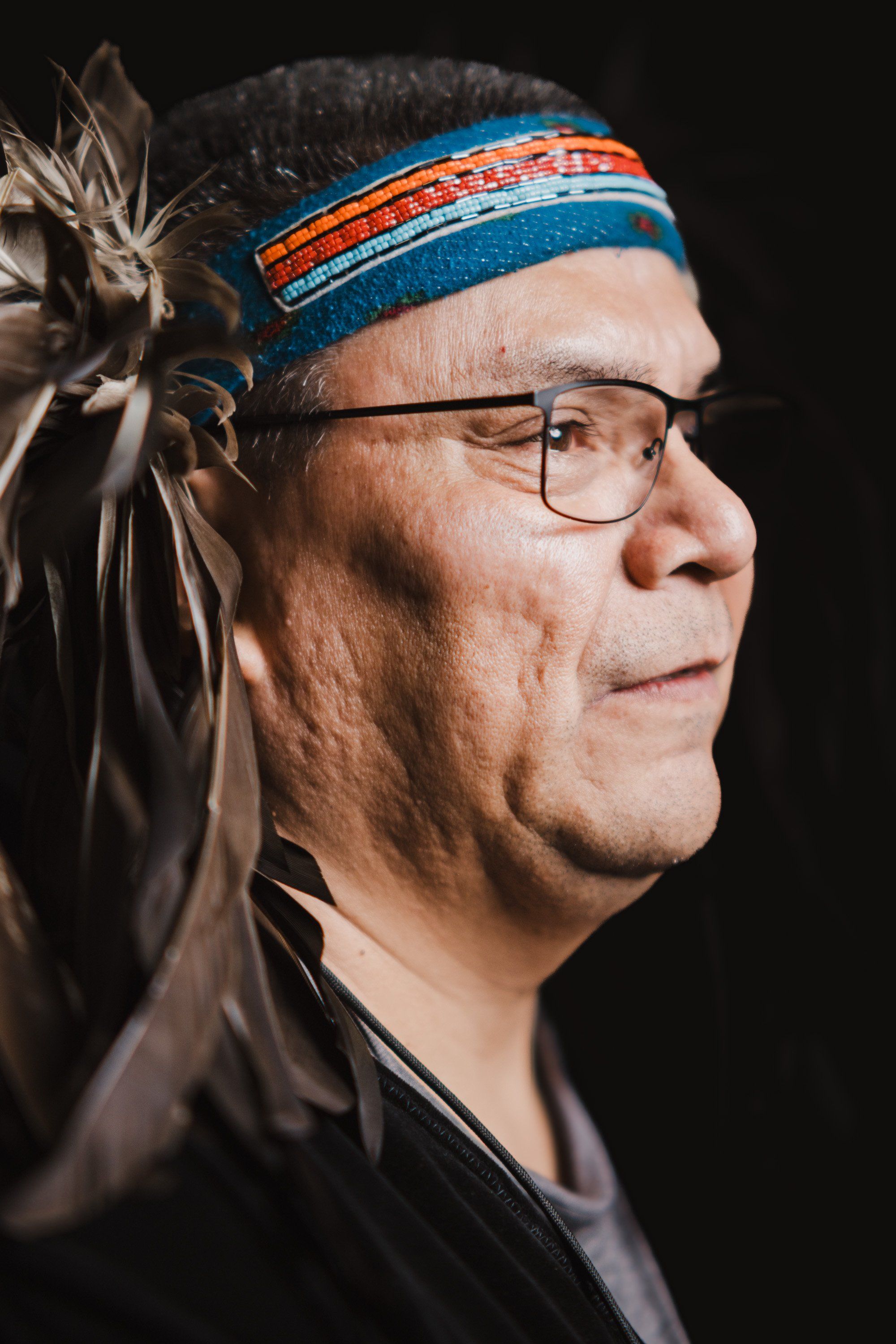 Indigenous man in profile wearing glasses, blue and orange headband, and traditional feathered regalia against black background