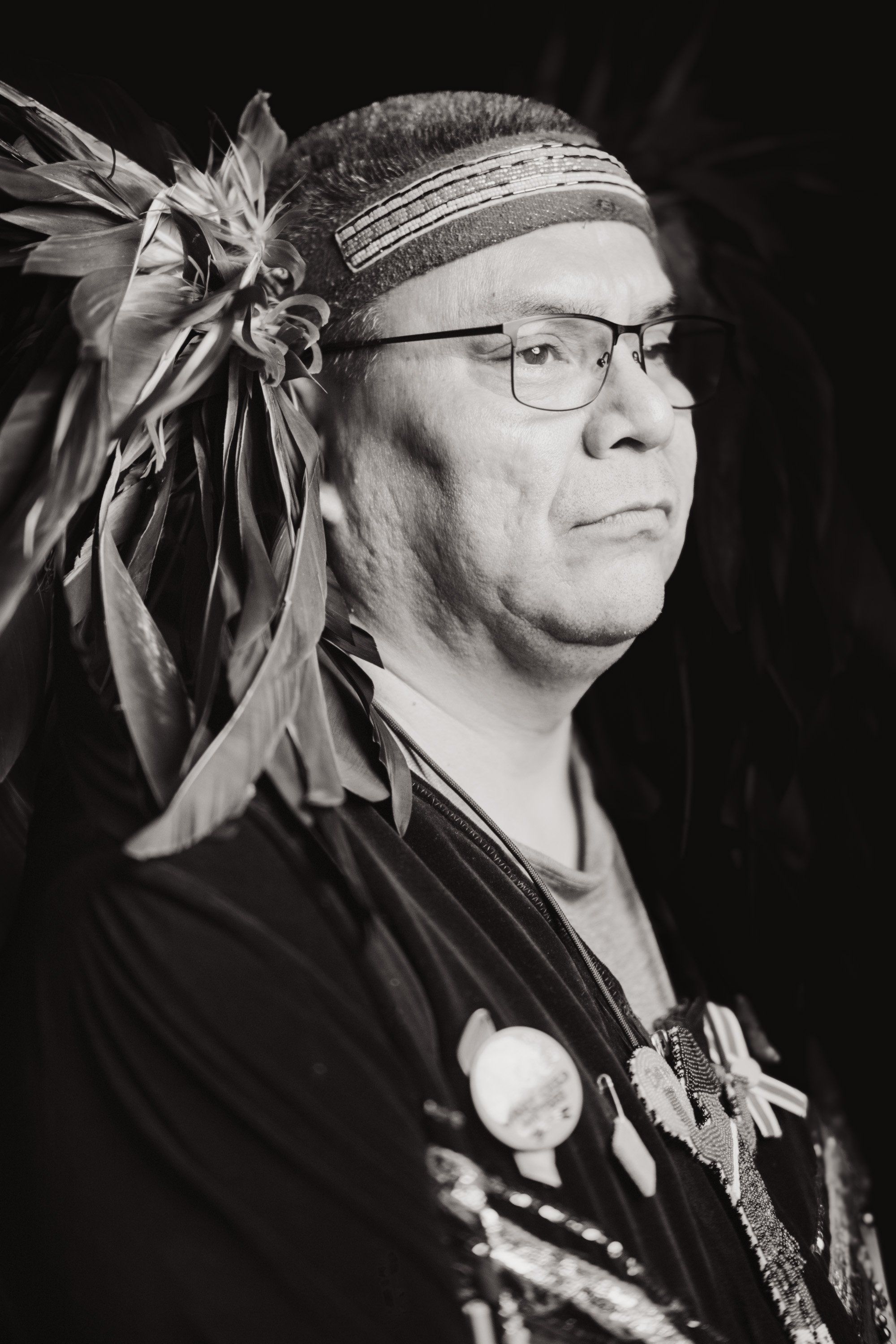 Man wearing glasses and headband with feathered ornament, shown in profile against dark background, wearing traditional regalia with pendant