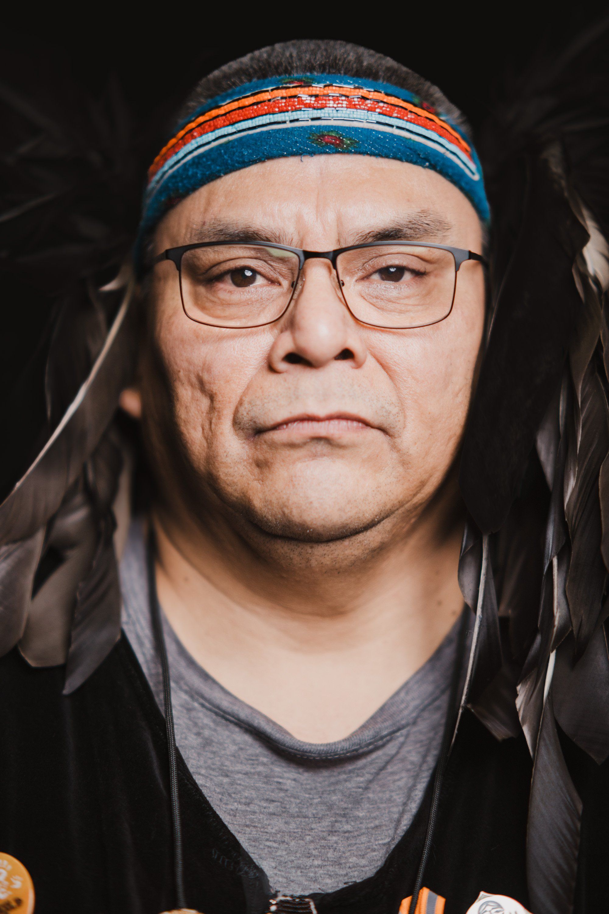 Portrait of a man wearing glasses and a colorful headband, with long dark hair, against a dark background