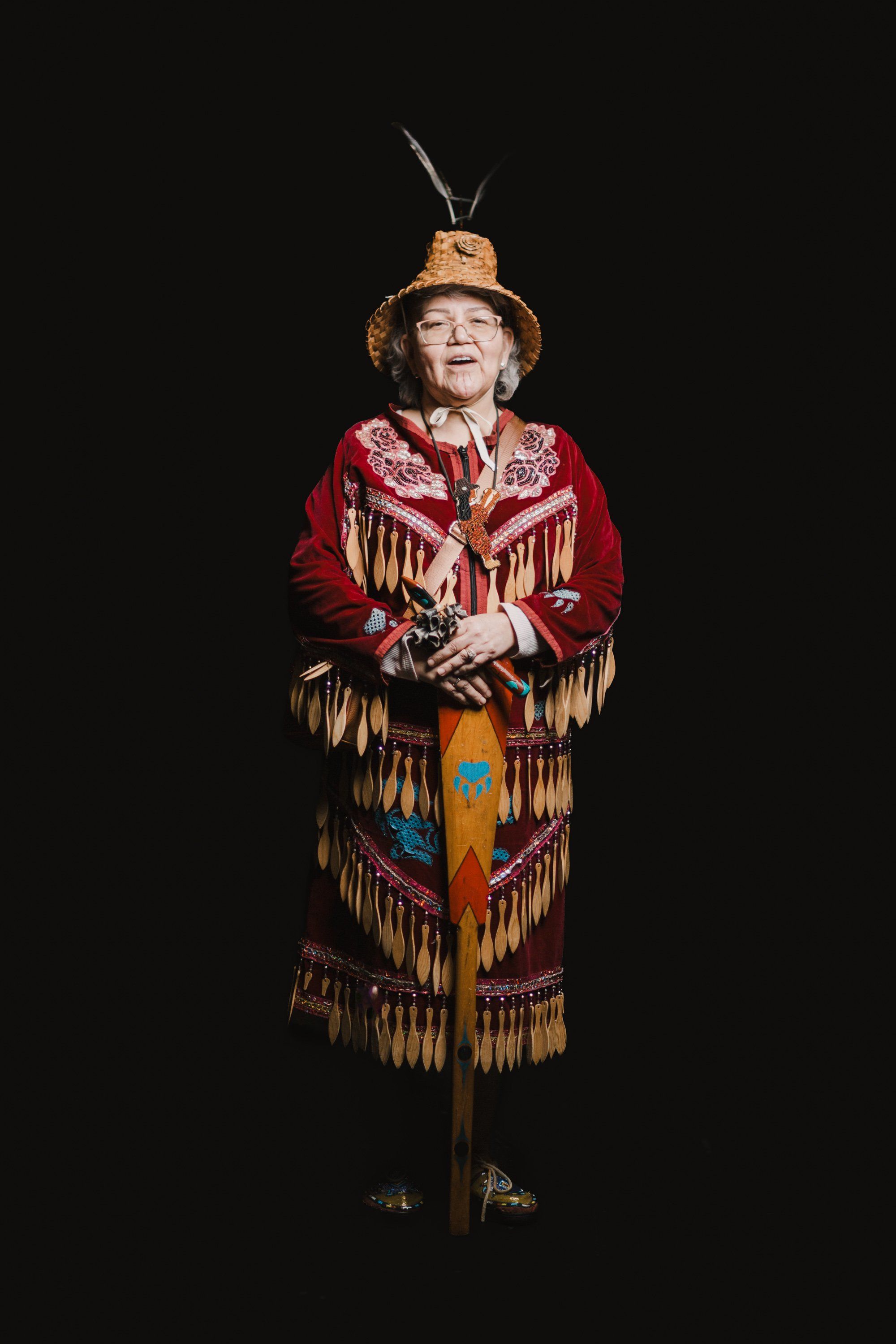 Elder in traditional red and gold regalia with fringe and ornate beadwork stands against black background, wearing a decorated hat with feathers