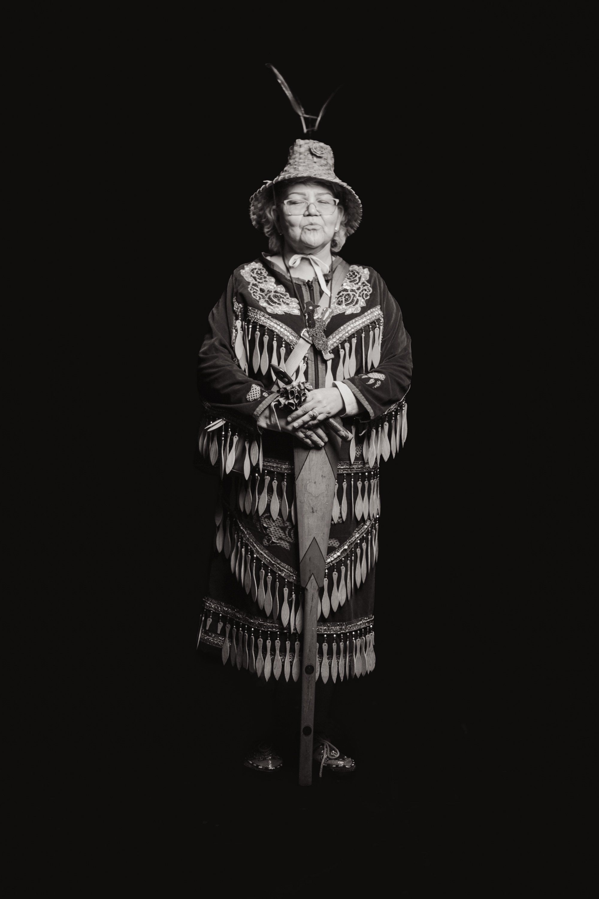Coast Salish performer in fringed regalia and feathered headdress stands against black background, hands clasped at chest