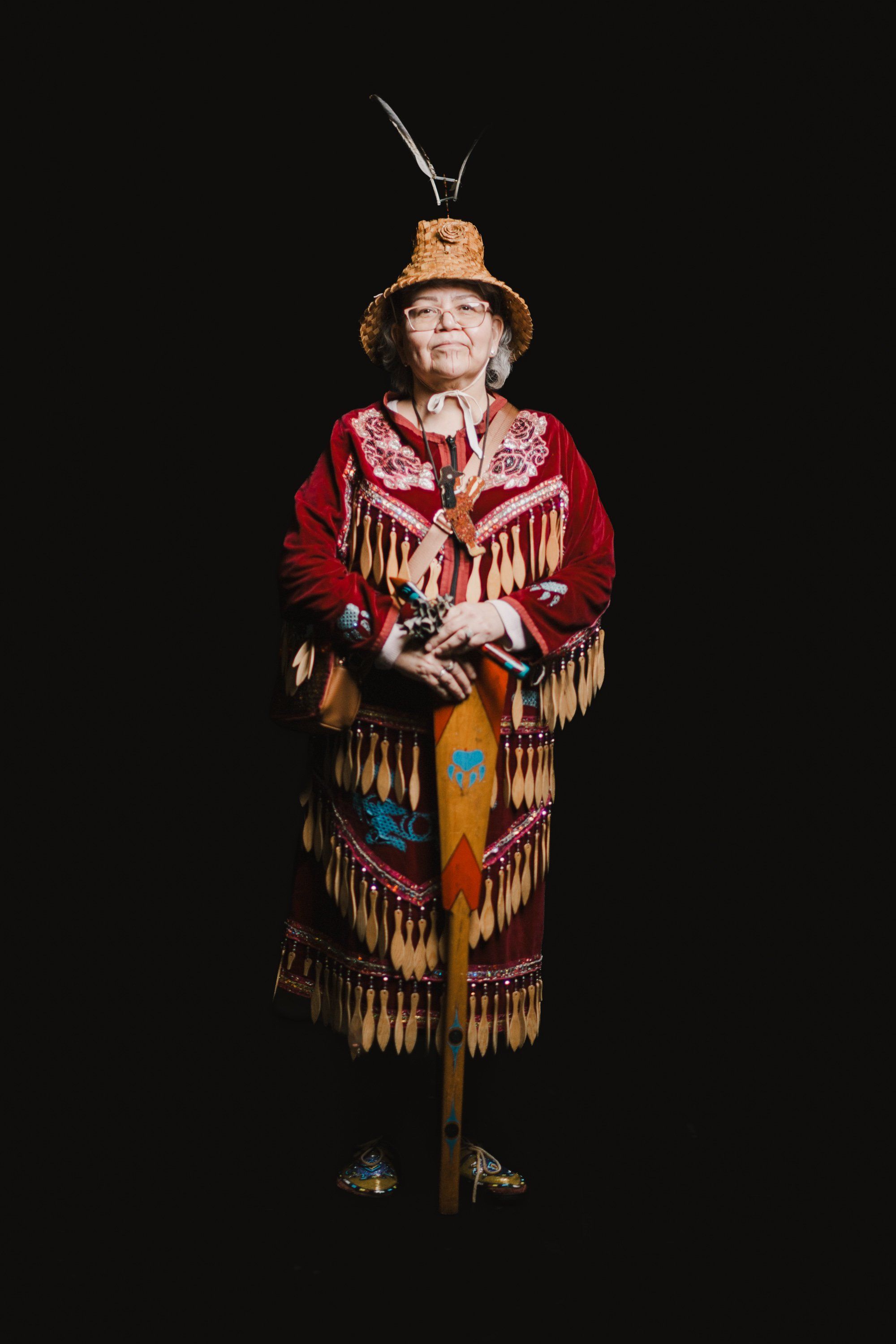 Elder in vibrant red and gold fringed regalia and woven hat stands against black background, holding staff with focused upward gaze