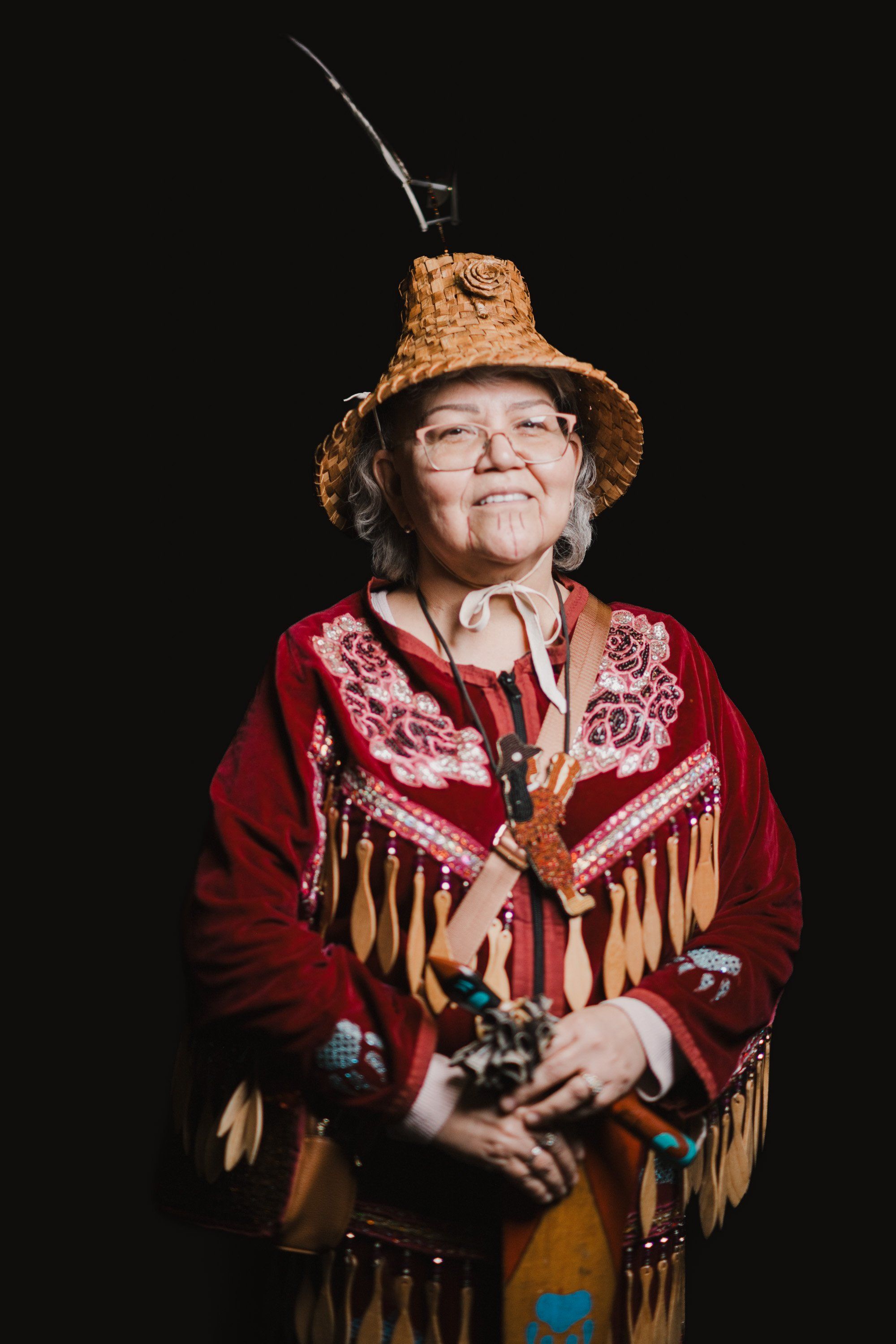 Elder in cork hat with feather and red fringed regalia smiles warmly against black background, holding traditional items