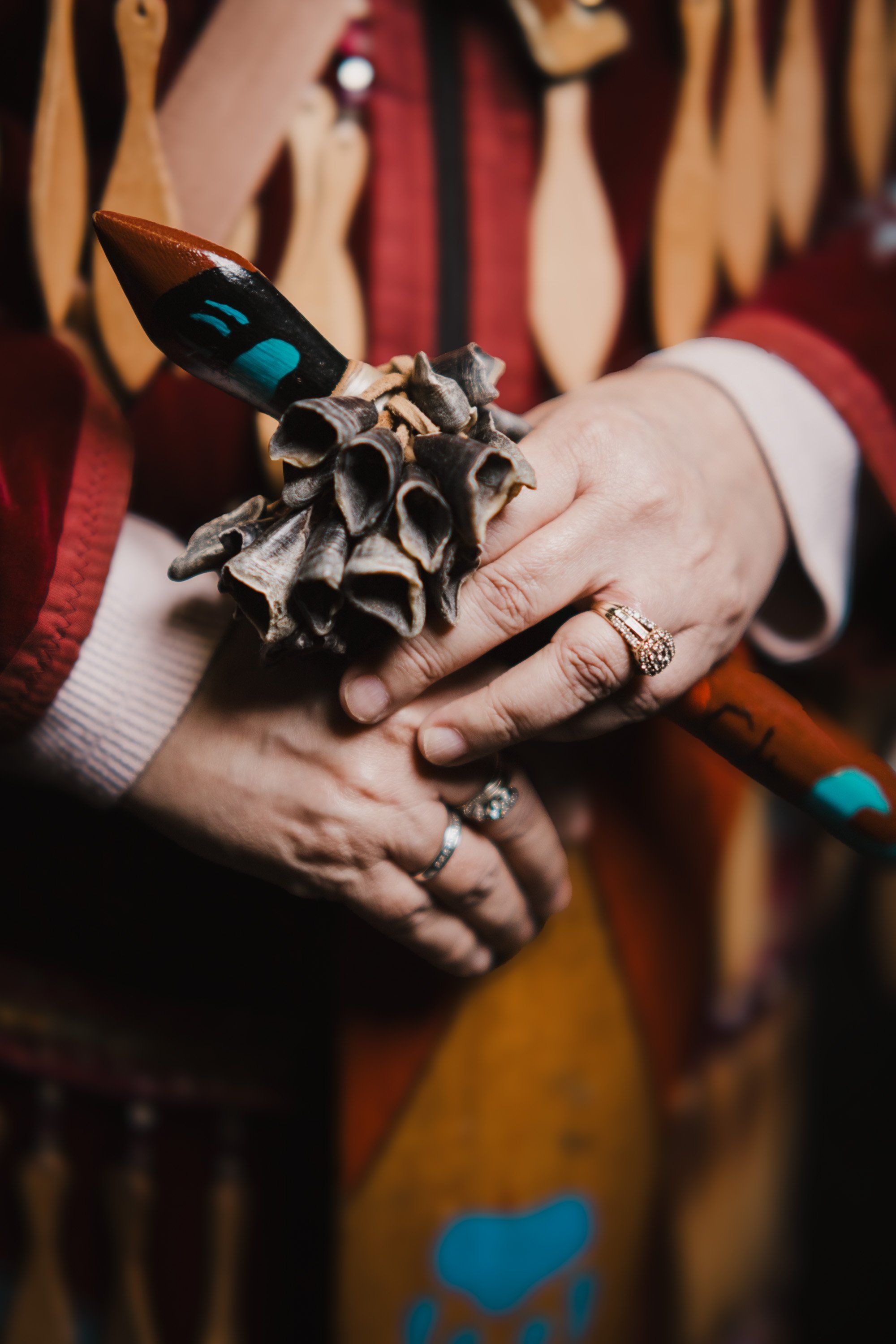 Hands adorned with silver rings hold a carved wooden raven with blue inlay against a backdrop of red and gold regalia