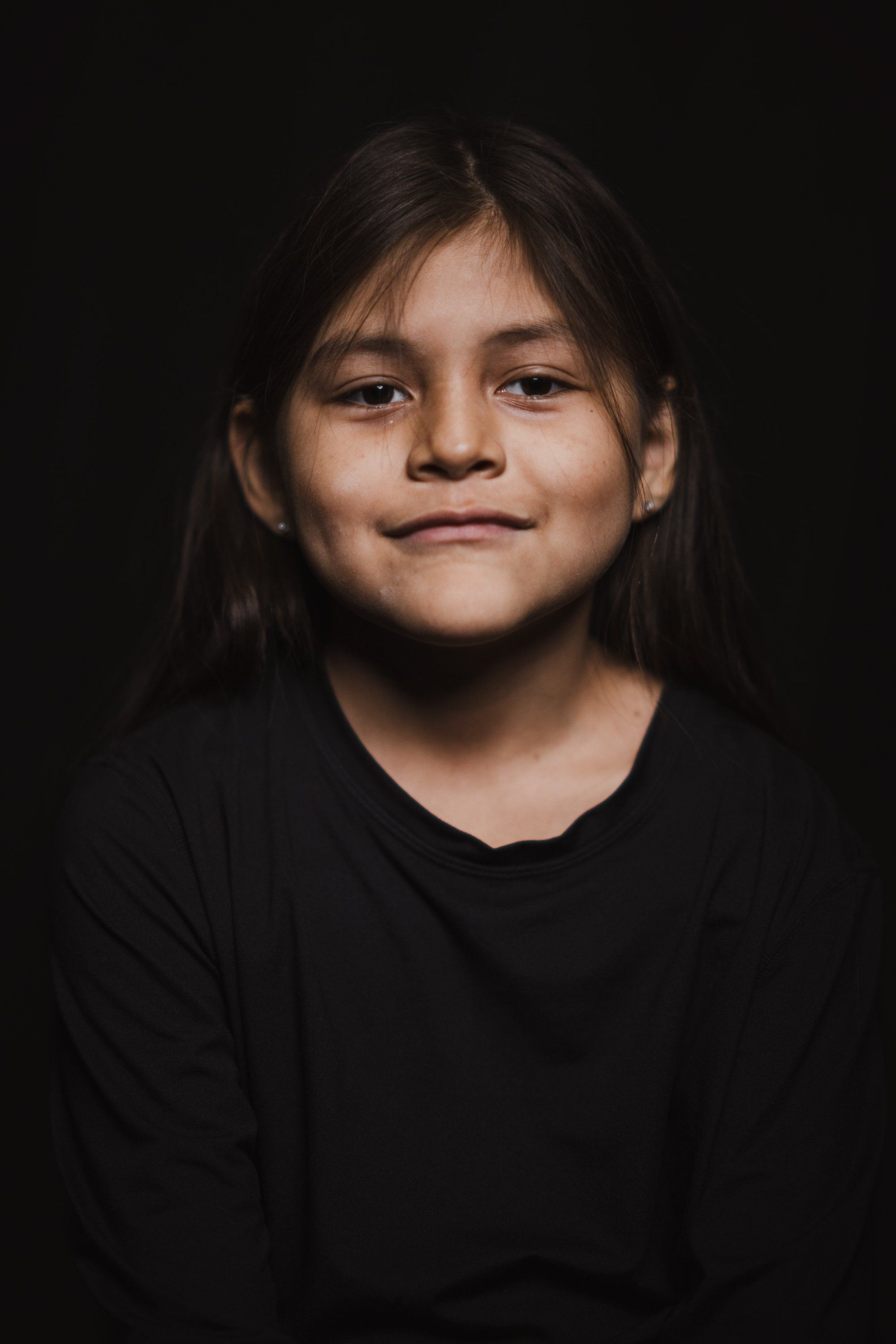 Young person with long dark hair wearing black against a dark background, gazing directly at camera with a gentle, confident expression