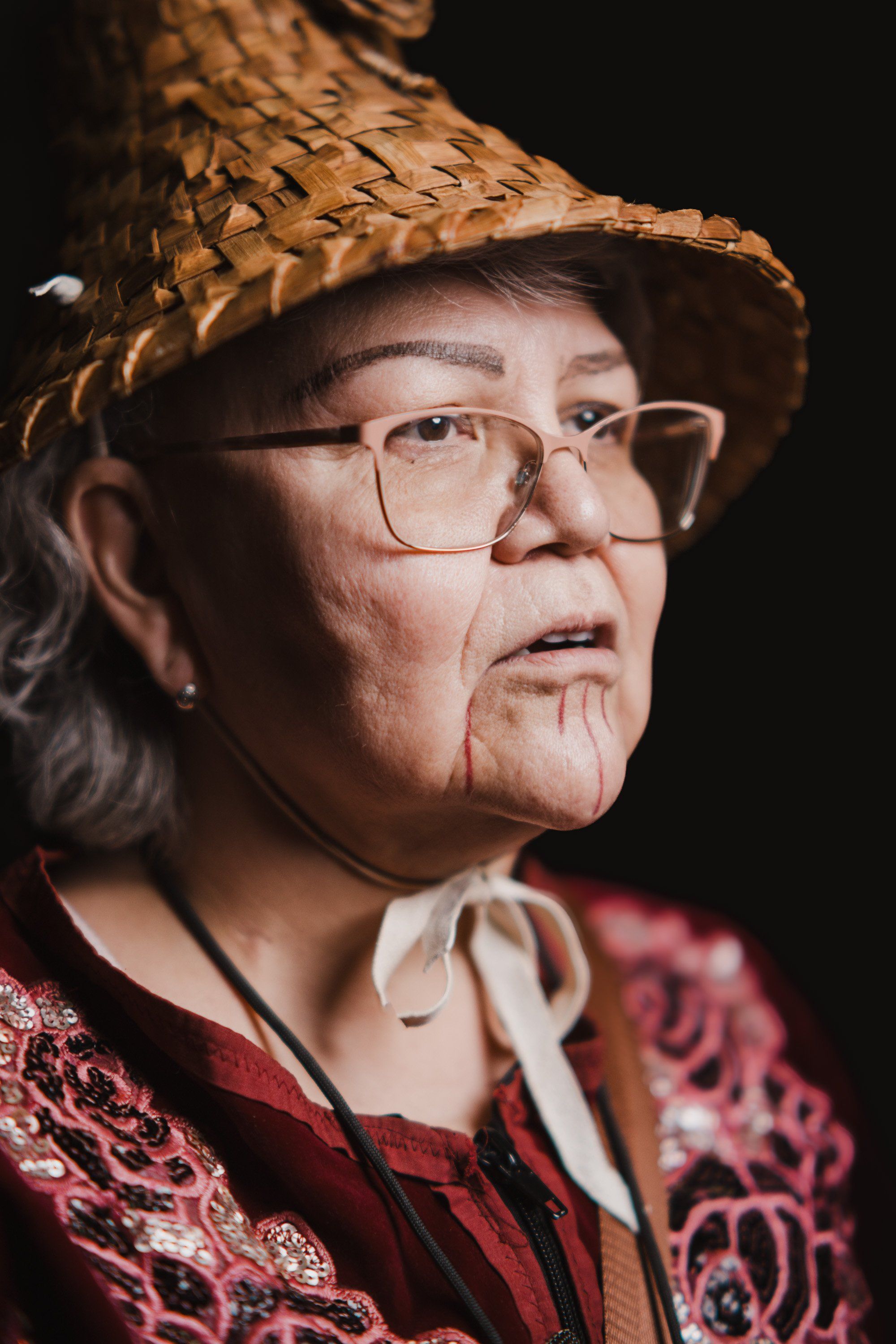 Elder in straw hat and glasses wearing red patterned regalia, photographed against dark background with warm side lighting