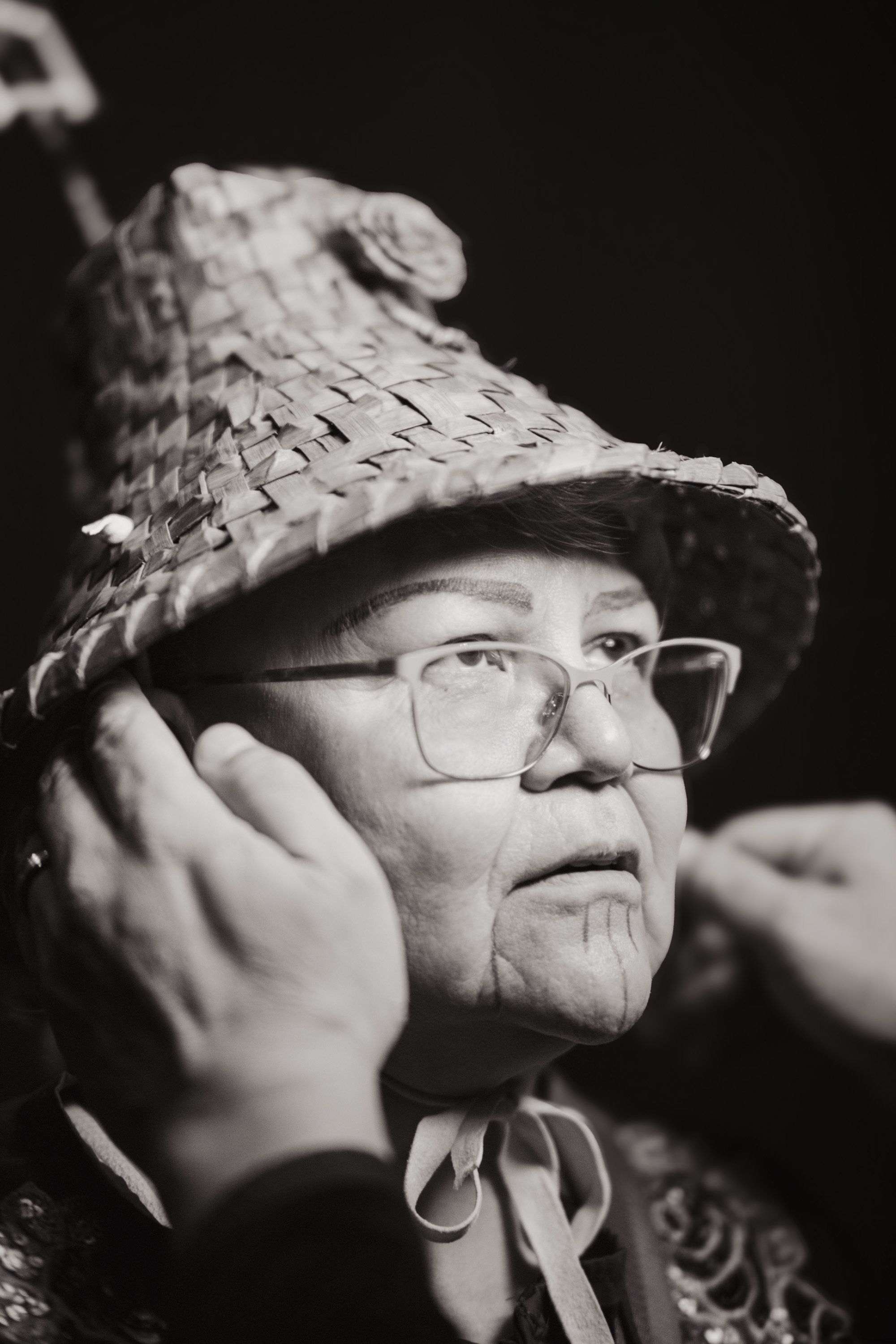 Elder wearing glasses and traditional woven hat gazes thoughtfully to the side, hands raised near face in contemplative pose against dark background