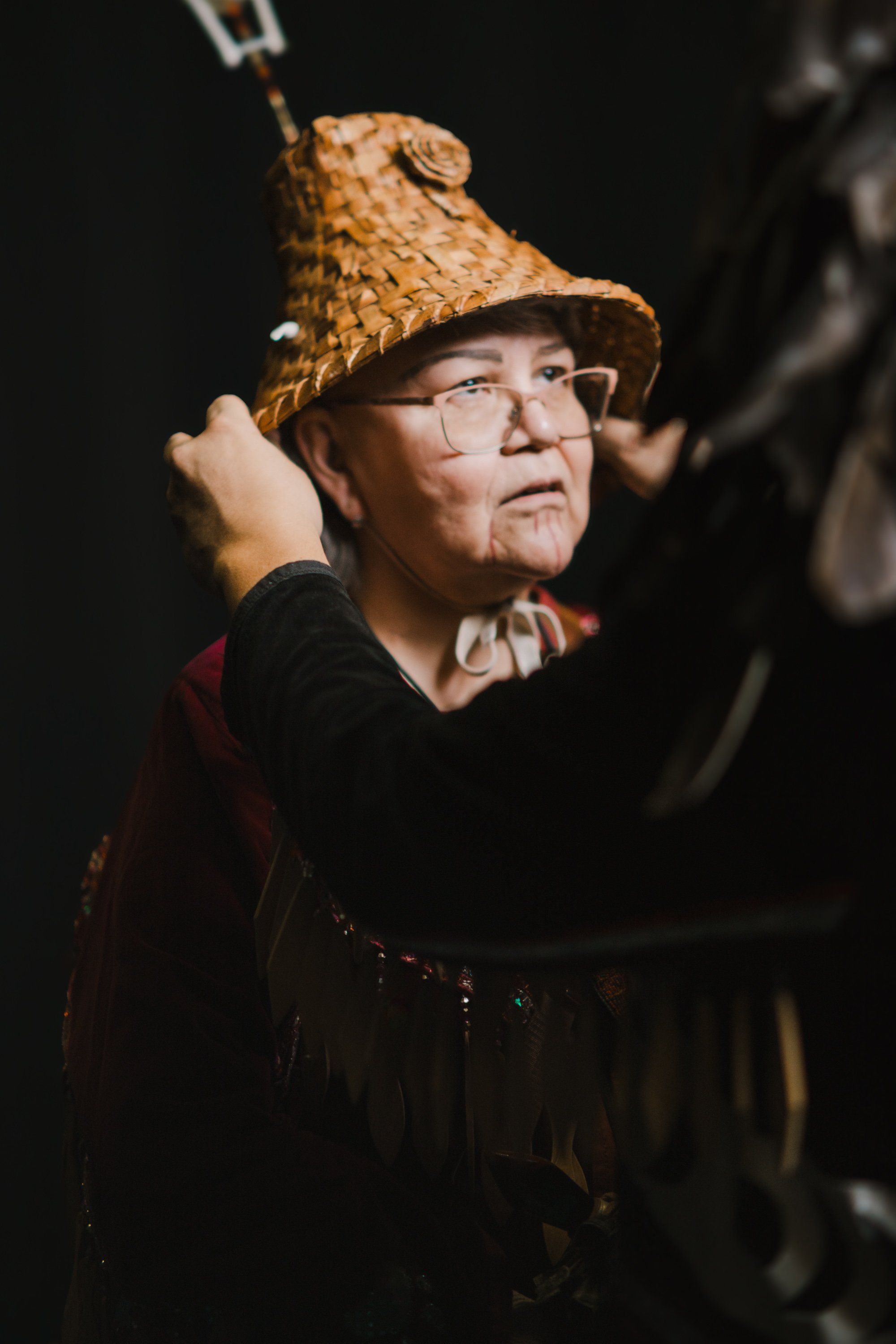 Indigenous performer in woven cedar hat and dark regalia gazes upward against black background, holding ceremonial staff