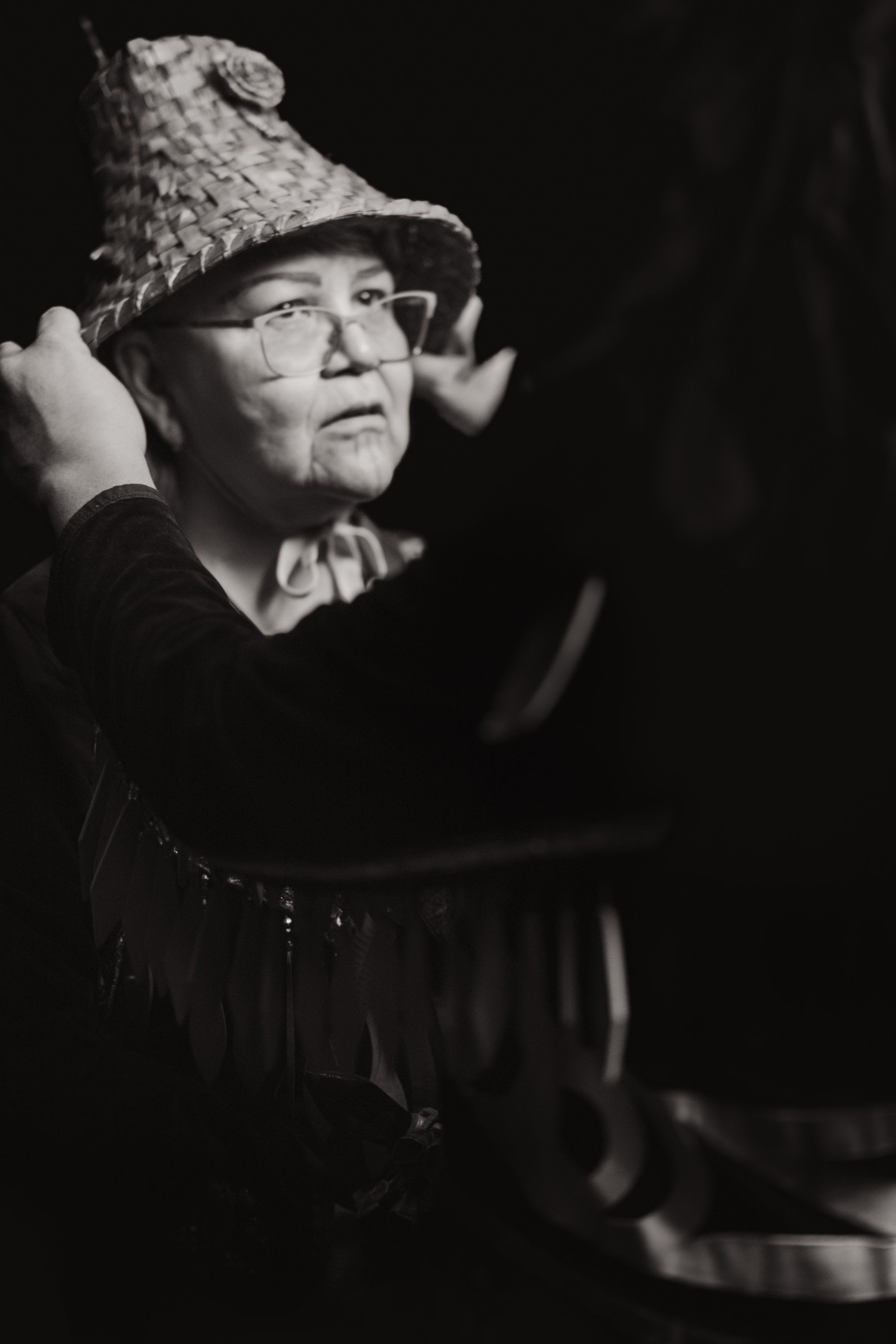 Performer in traditional Coast Salish regalia and glasses, dramatic side-lit black and white portrait against dark background