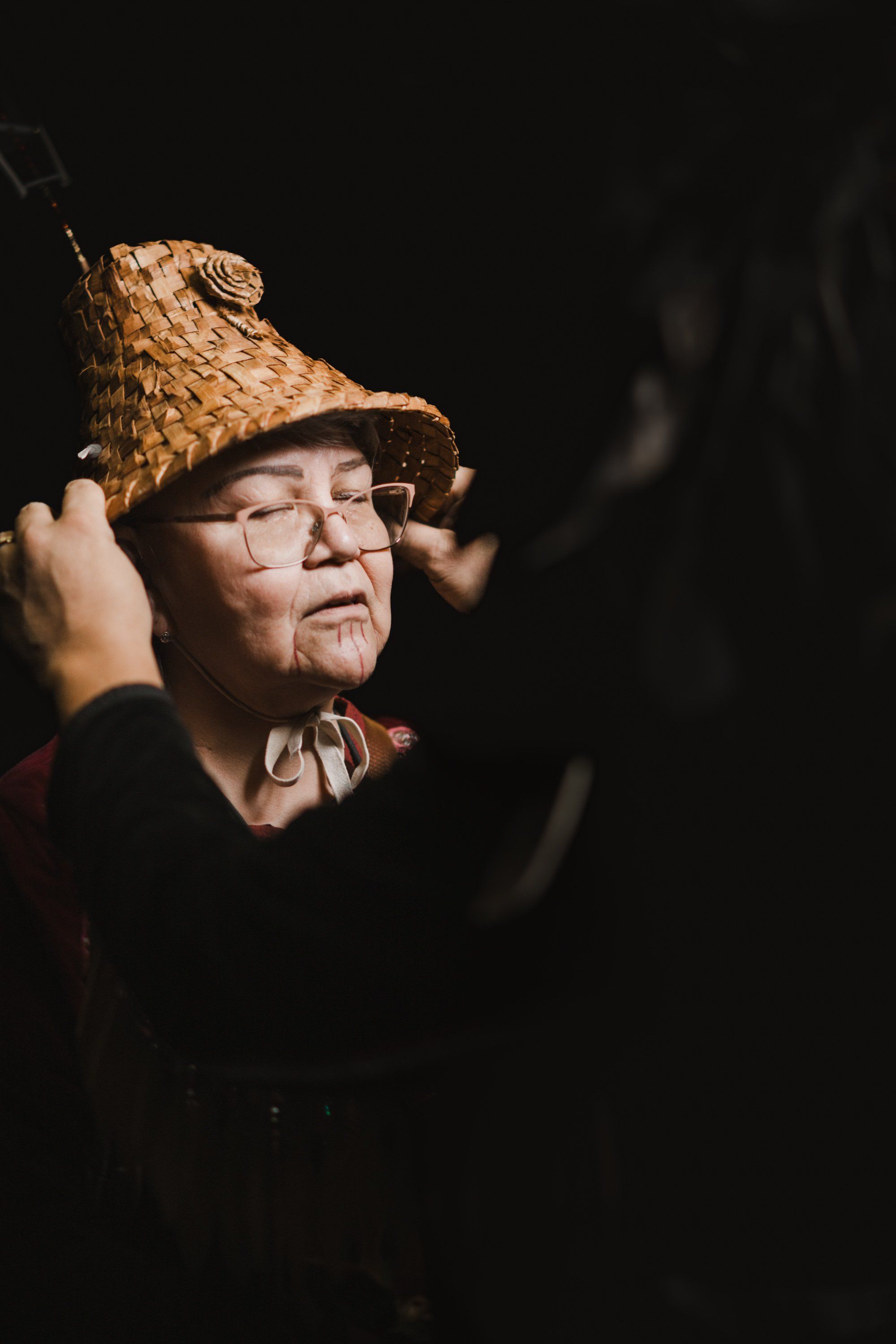 Elder in traditional woven hat and glasses, eyes closed in serene expression, dramatically lit against dark background
