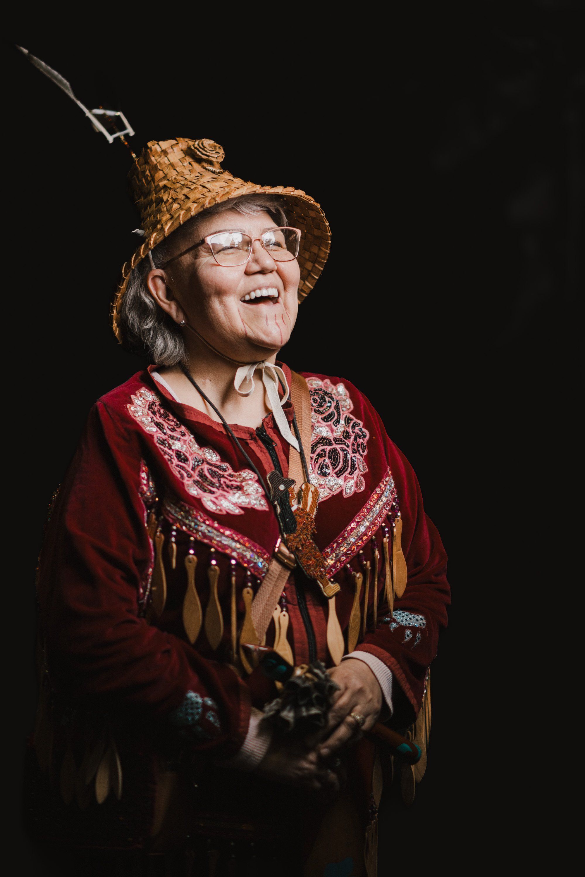 Elder in traditional Coast Salish regalia smiles upward against black background, wearing woven hat, burgundy blanket with fringe, and glasses