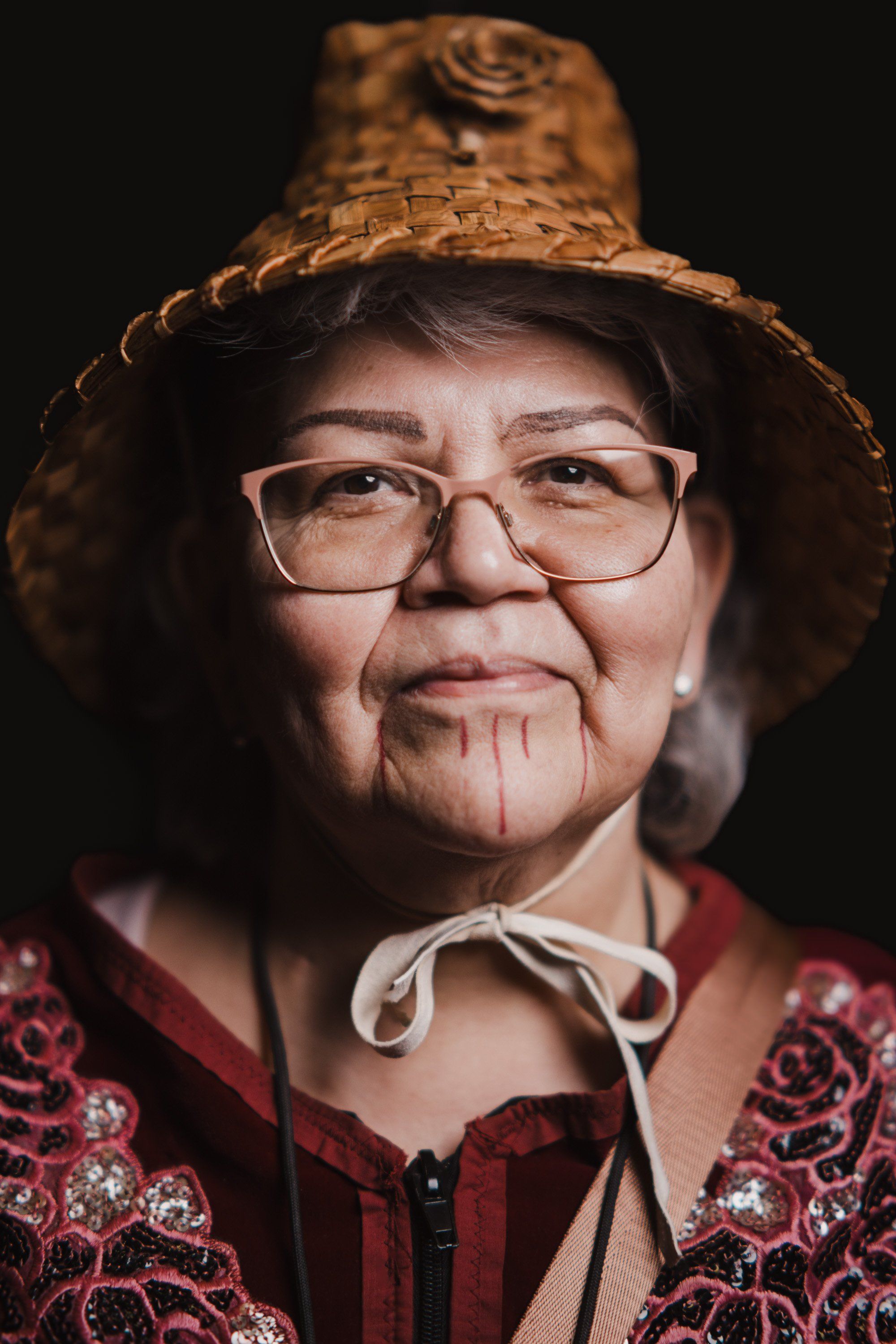 Elder in glasses and straw hat wearing burgundy beaded regalia smiles warmly against dark background