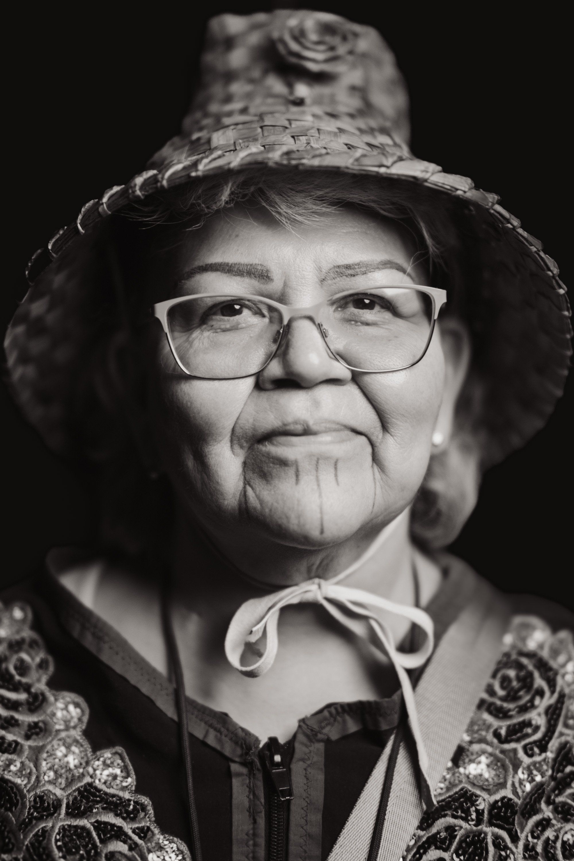 Portrait of an elder wearing glasses and a textured hat with chin strap, adorned with decorative regalia, against a dark background