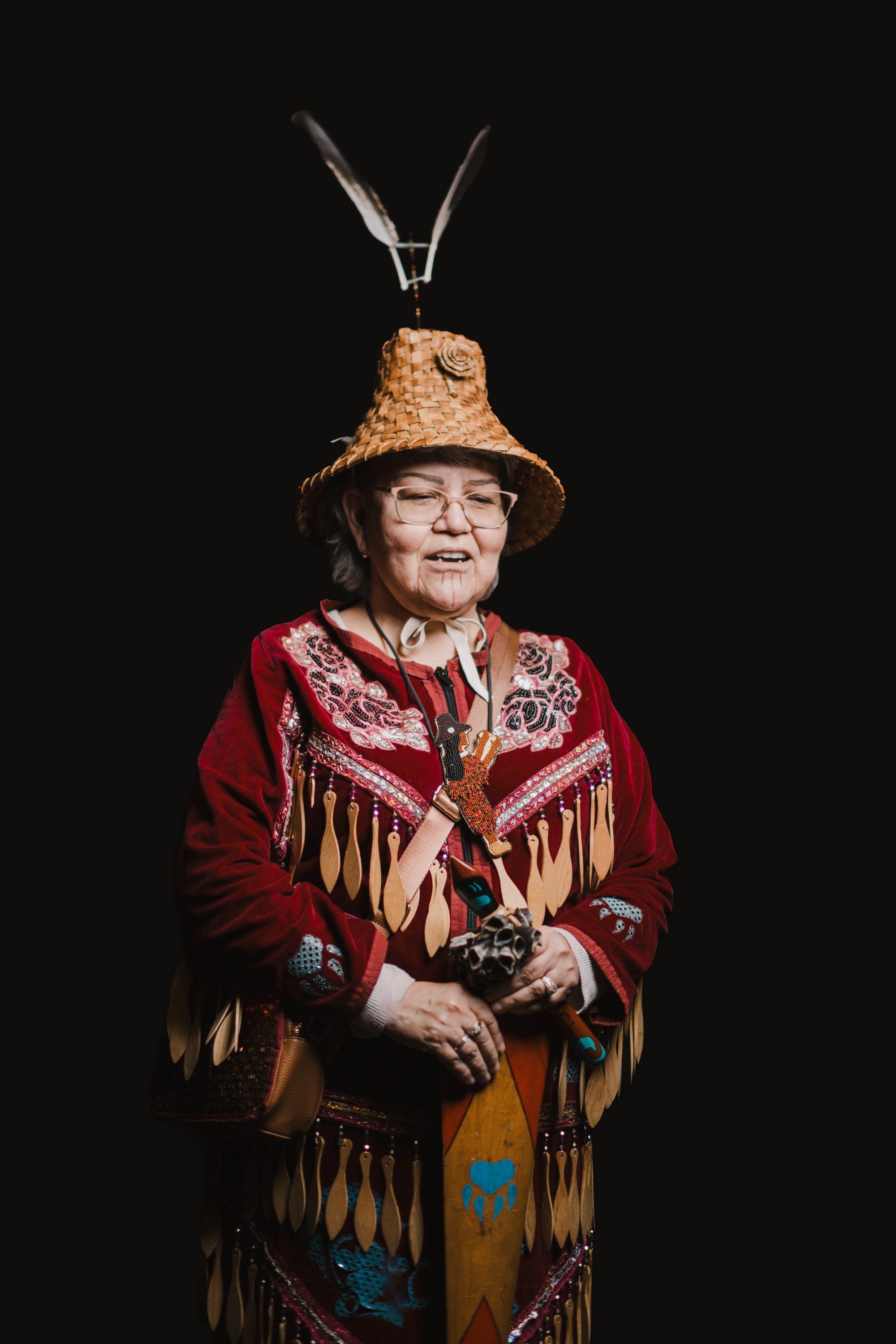 Elder in traditional red regalia and woven hat adorned with antlers, smiling warmly against dark background