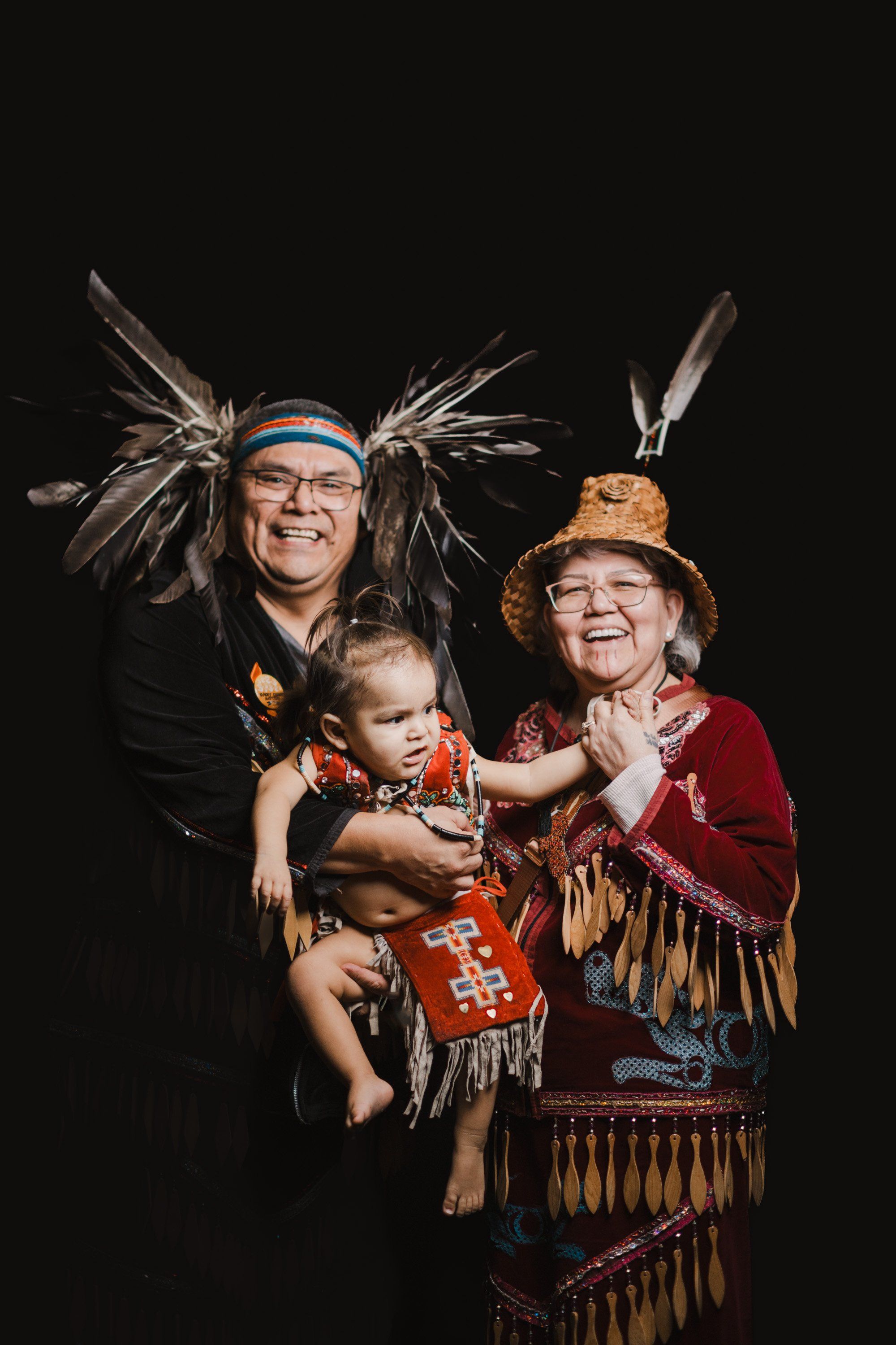 Three generations in regalia smile together against black backdrop, feathered headdresses and fringed clothing celebrating Coast Salish heritage