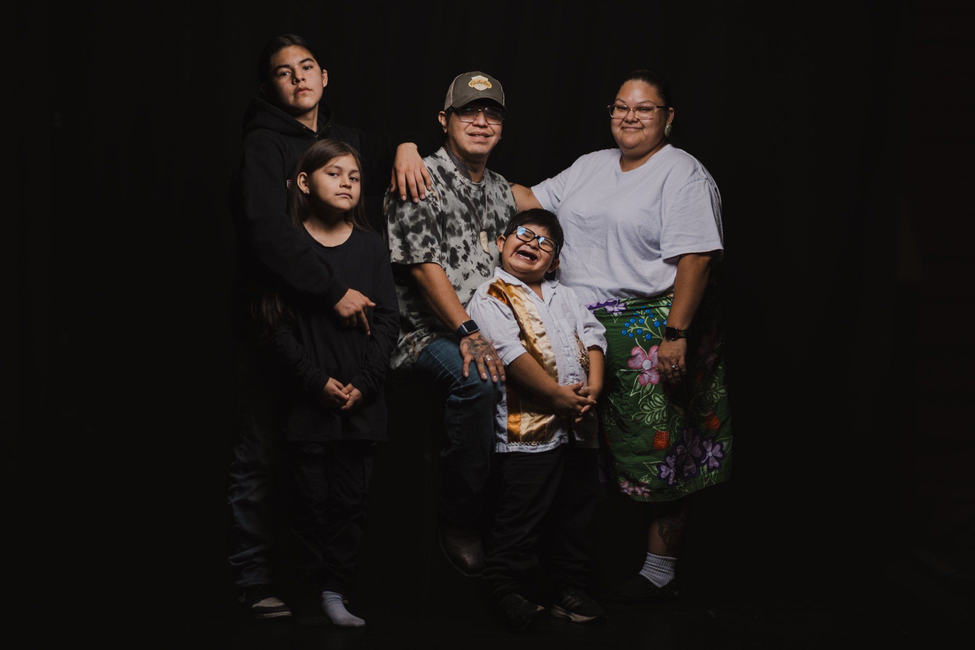 Five family members pose closely together against a black background, wearing dark and patterned clothing, their warm connection evident in their embraces an…