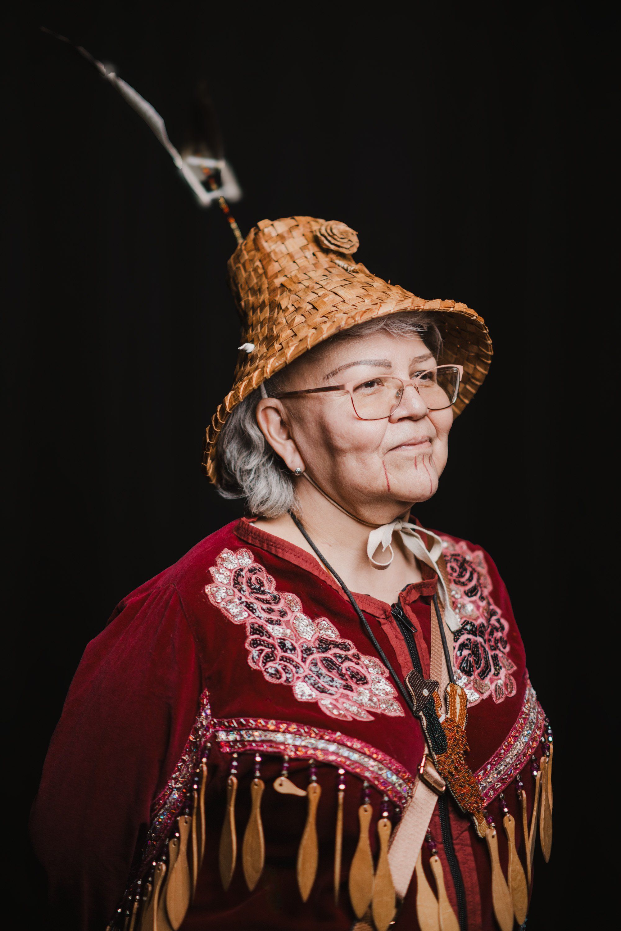 Elder in woven cedar hat and burgundy regalia with fringe and beadwork, wearing glasses, photographed against black background