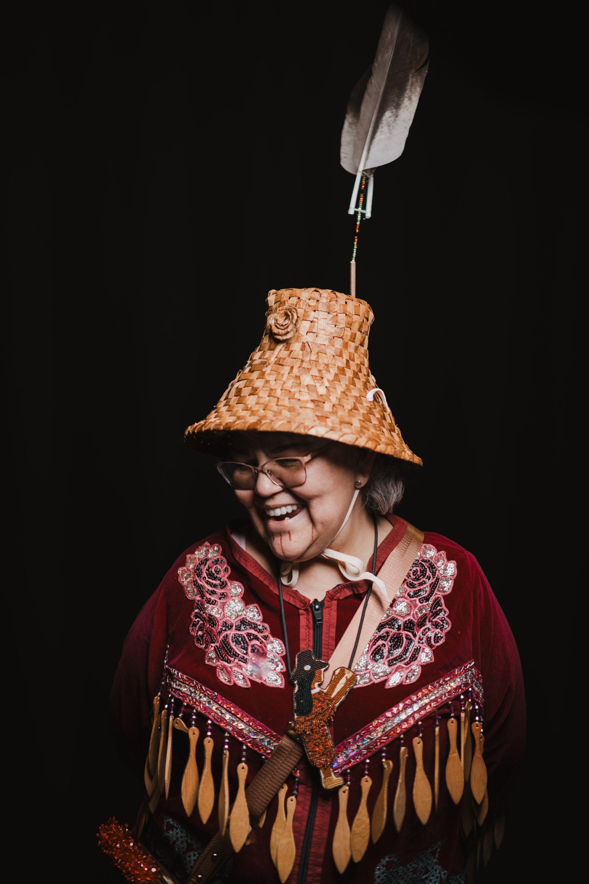 Elder in woven cedar hat and burgundy regalia with golden fringe smiles brightly against black background, feather suspended above
