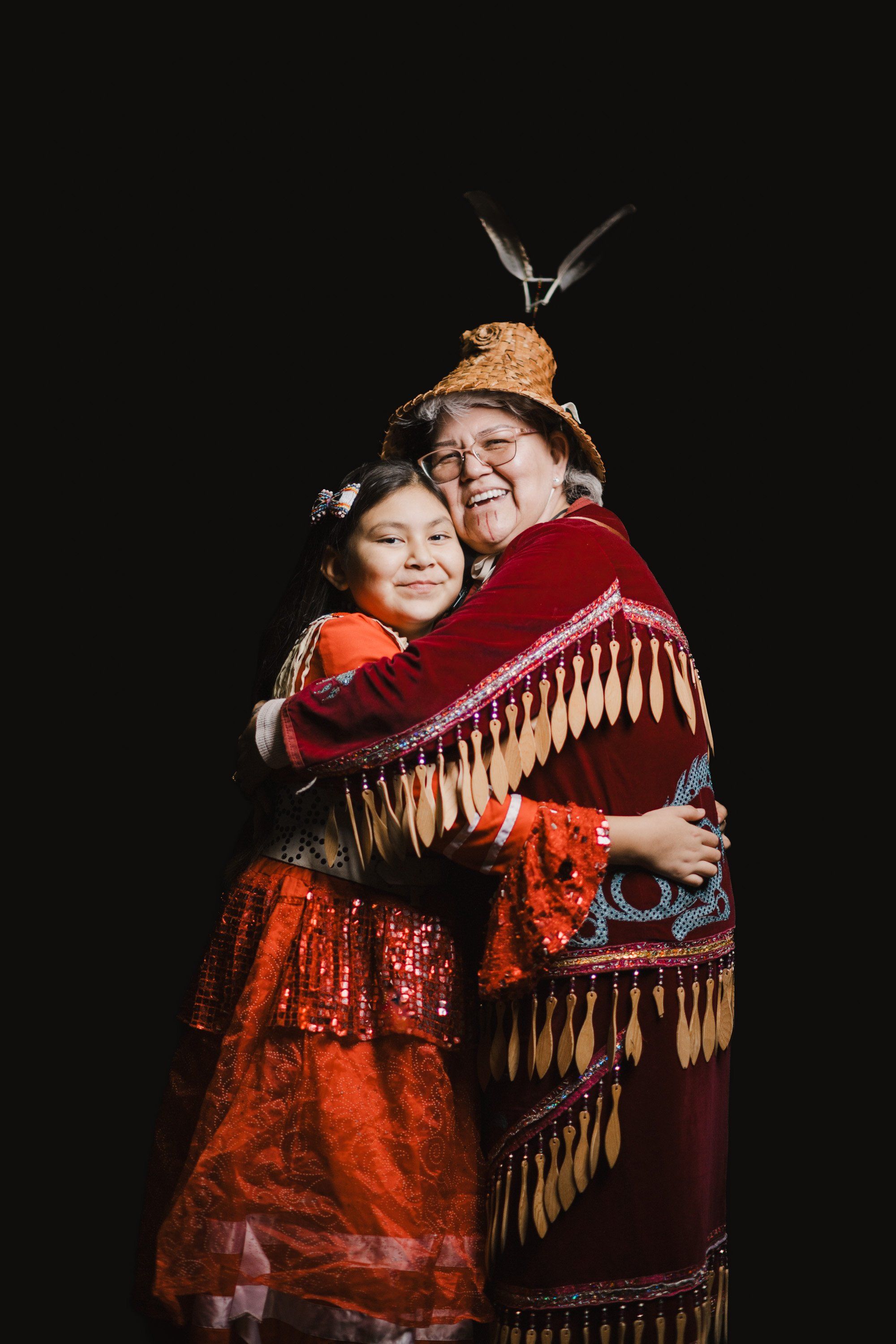 Two performers in vibrant traditional regalia embrace against a black background, wearing red and orange beaded clothing with fringed details and woven hats