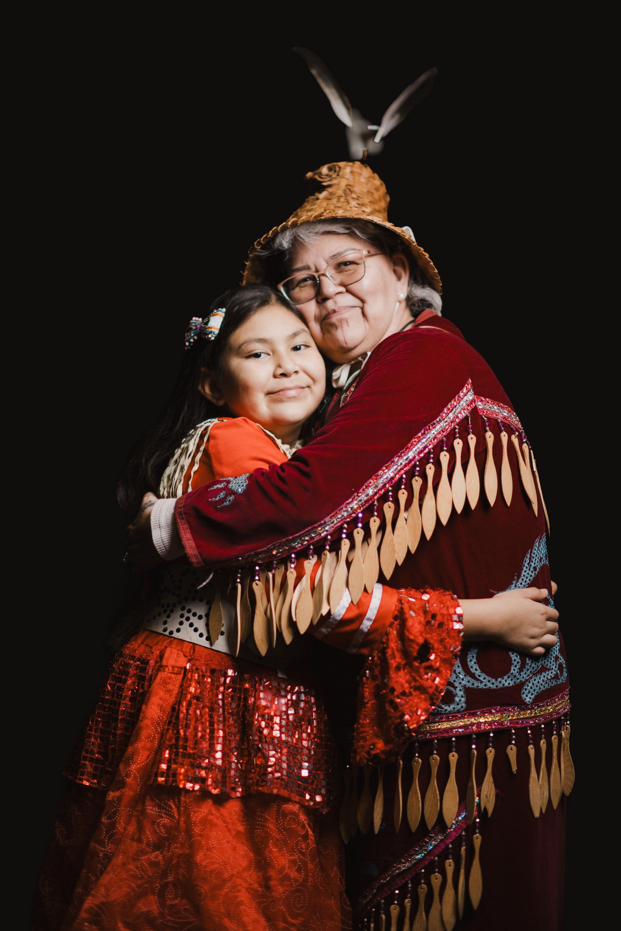 Two people in vibrant red and orange regalia with beaded details embrace against a black background, a carved bird pendant suspended above them