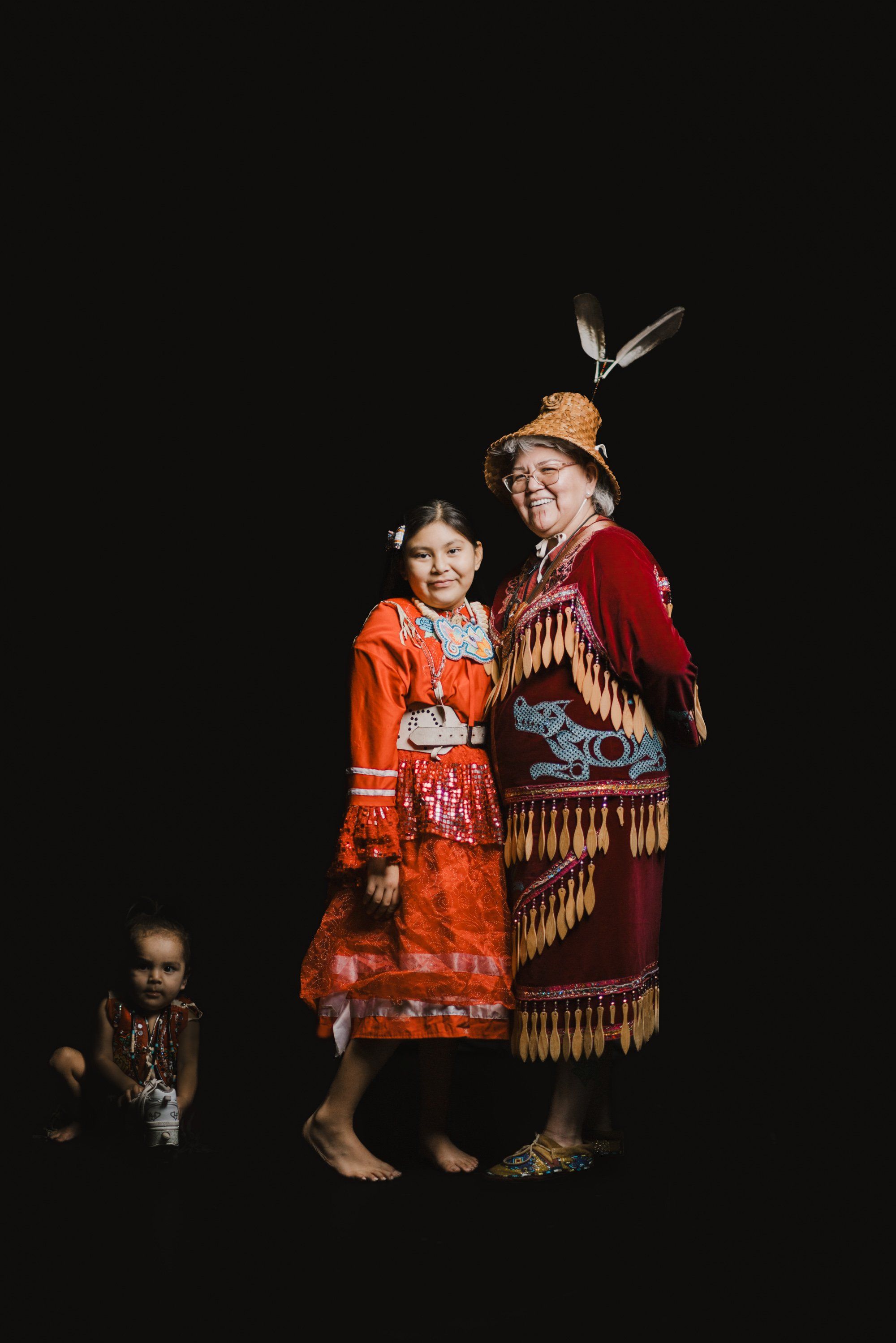 Three generations in Coast Salish regalia stand against black, the elder and child in vibrant red and orange textiles, feathers adorning their hair