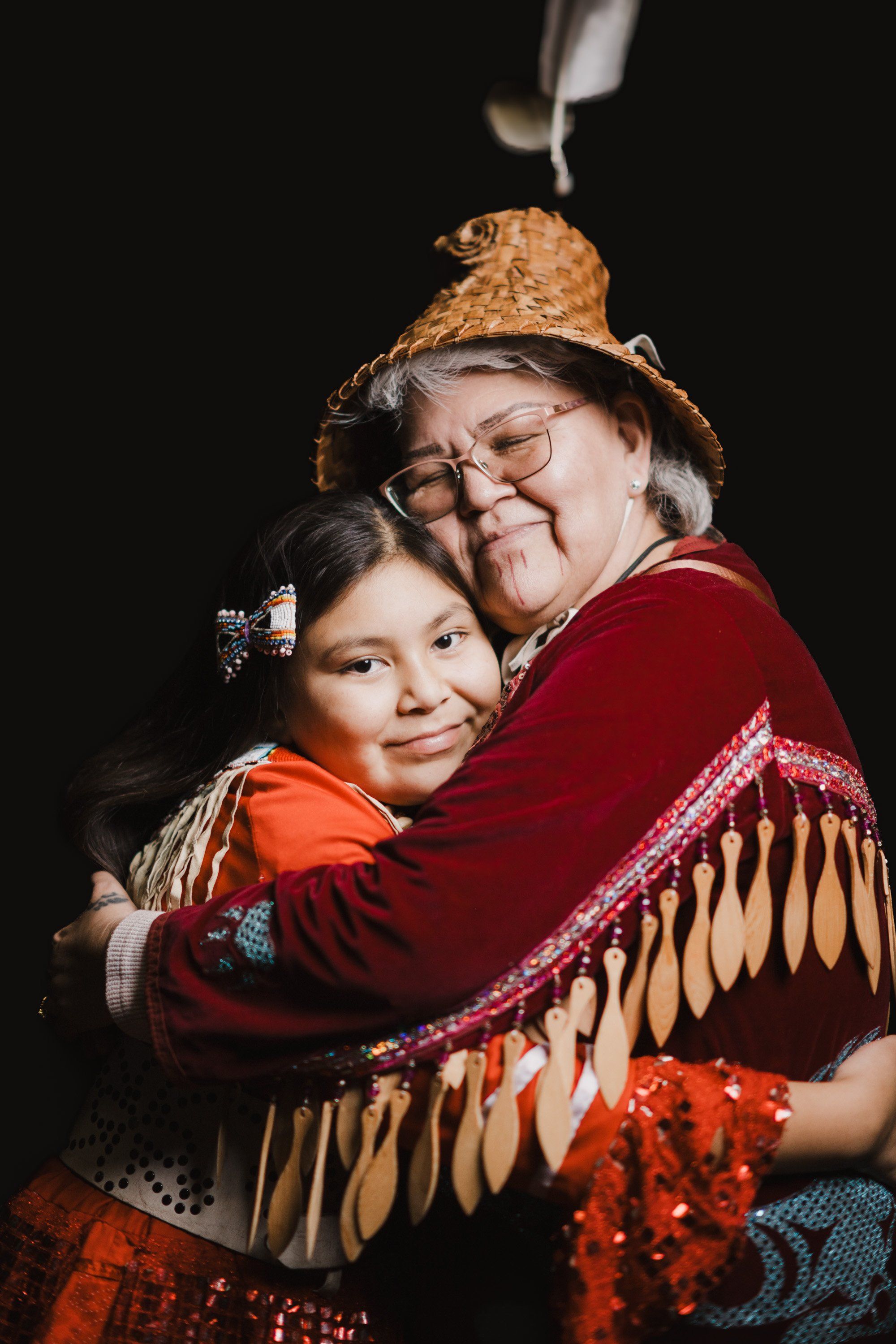 Elder and child embrace warmly in traditional regalia against black, their genuine joy illuminated by soft studio light