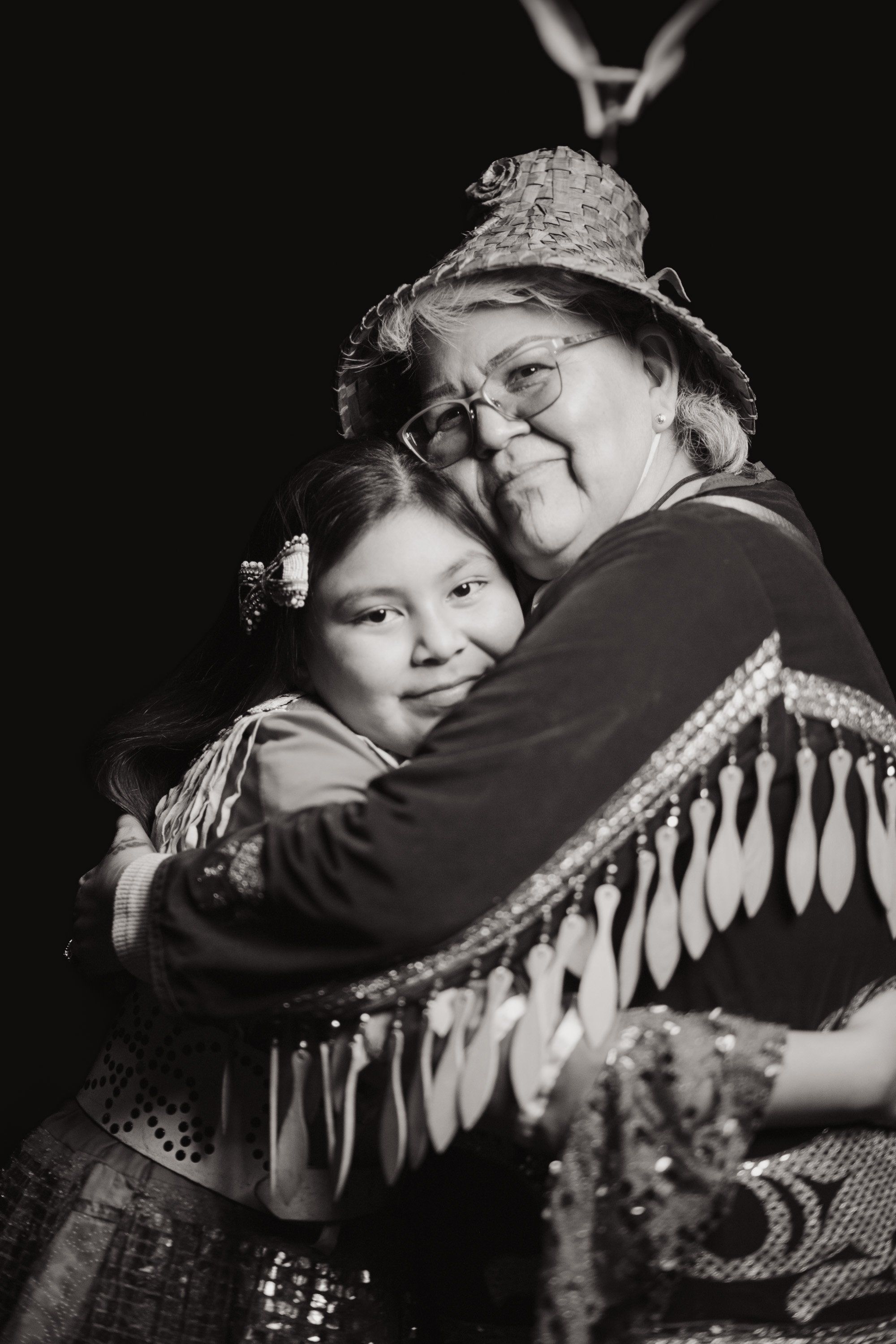 Two figures embrace warmly against a dark background, one wearing glasses and a decorative hat with fringe shawl, the other in traditional regalia, sharing a…